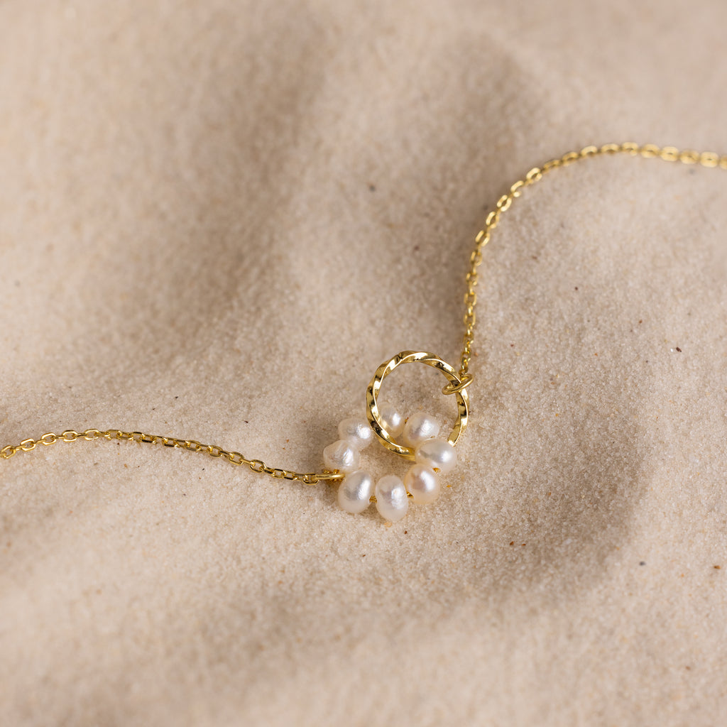Interlocking Pearl Necklace featuring a gold chain with a circular twisted ring charm and delicate pearl accents, shown elegantly on soft sand.