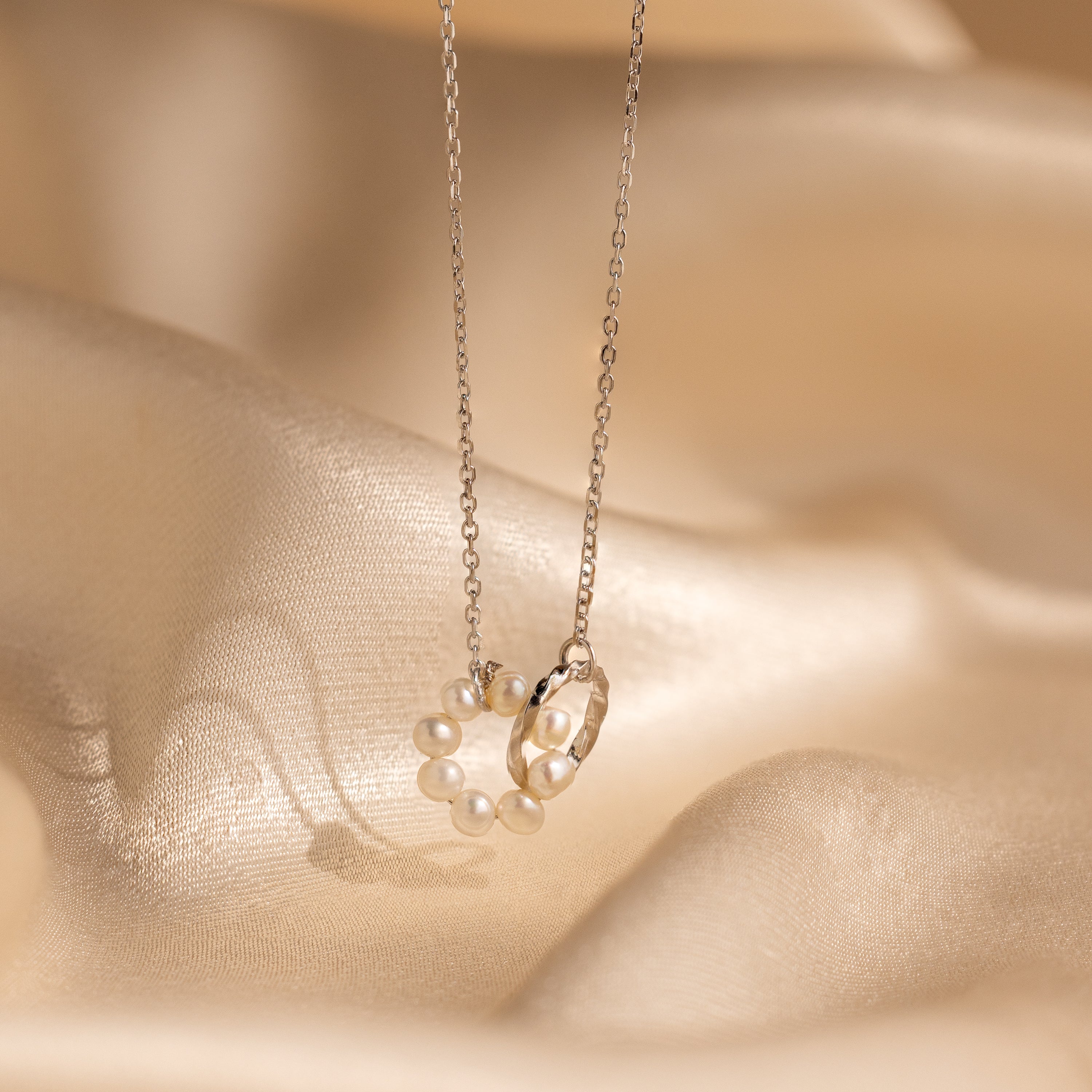 The Interlocking Pearl Necklace in Silver features pearl accents and a twisted ring charm, elegantly displayed on soft, beige satin fabric.