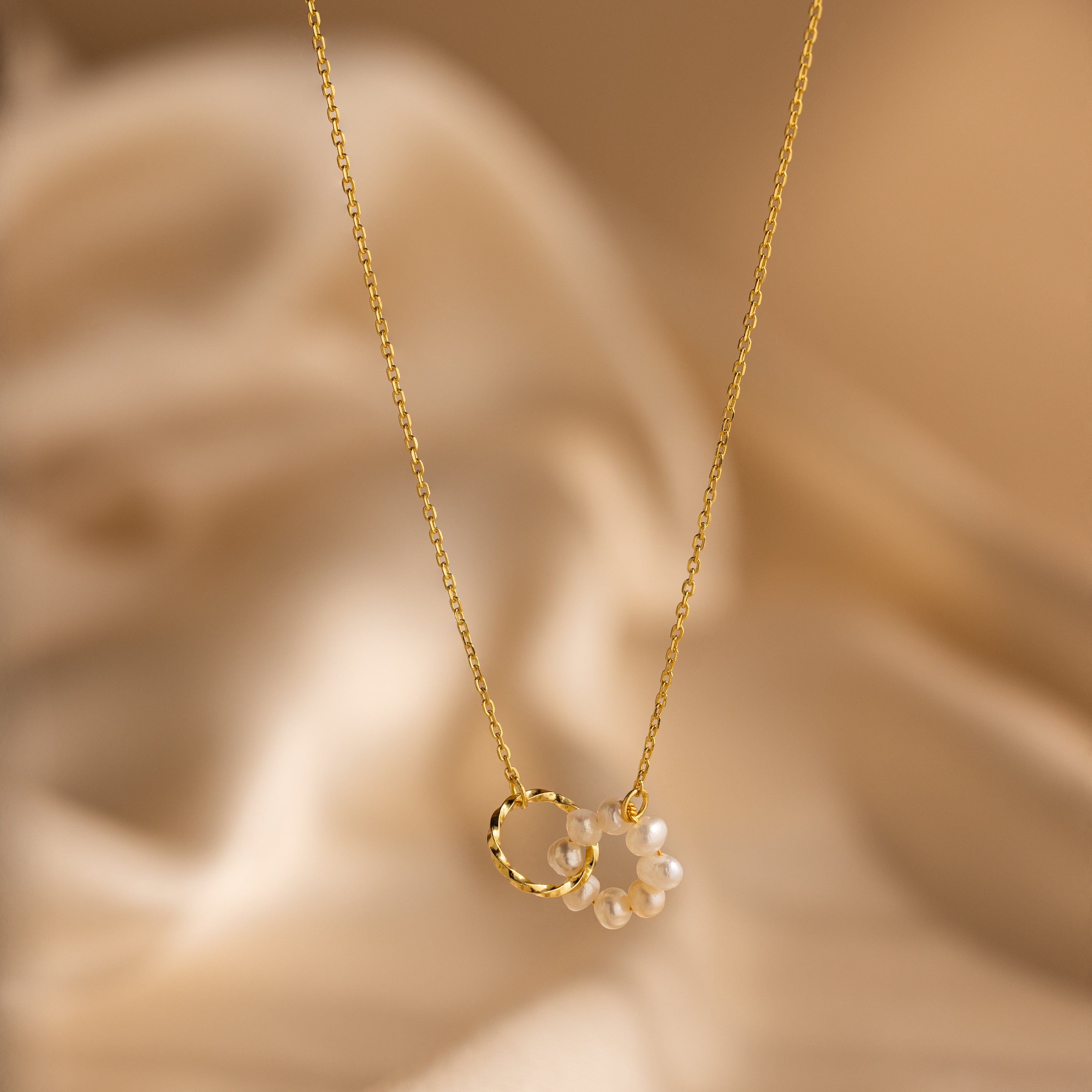 The Interlocking Pearl Necklace features a gold chain with a twisted ring charm and small white beads, elegantly displayed on beige fabric—perfect for any occasion.