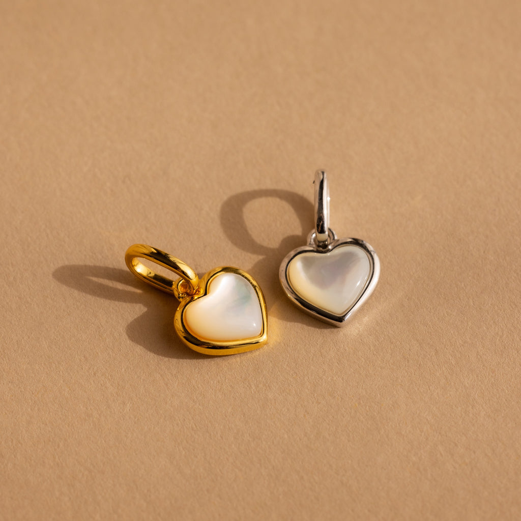 The Mother of Pearl Heart Charm in Sterling Silver features a heart-shaped pendant with a white inlay inspired by vintage jewelry, adding timeless elegance to any look. Set against a beige background.