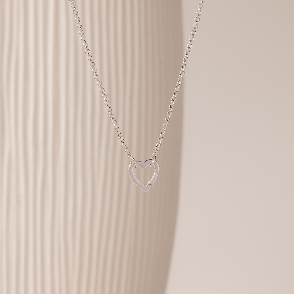 The Rolo Heart Clasp Necklace in White Gold features a delicate open heart pendant on a classic rolo chain, shown against a neutral textured background.