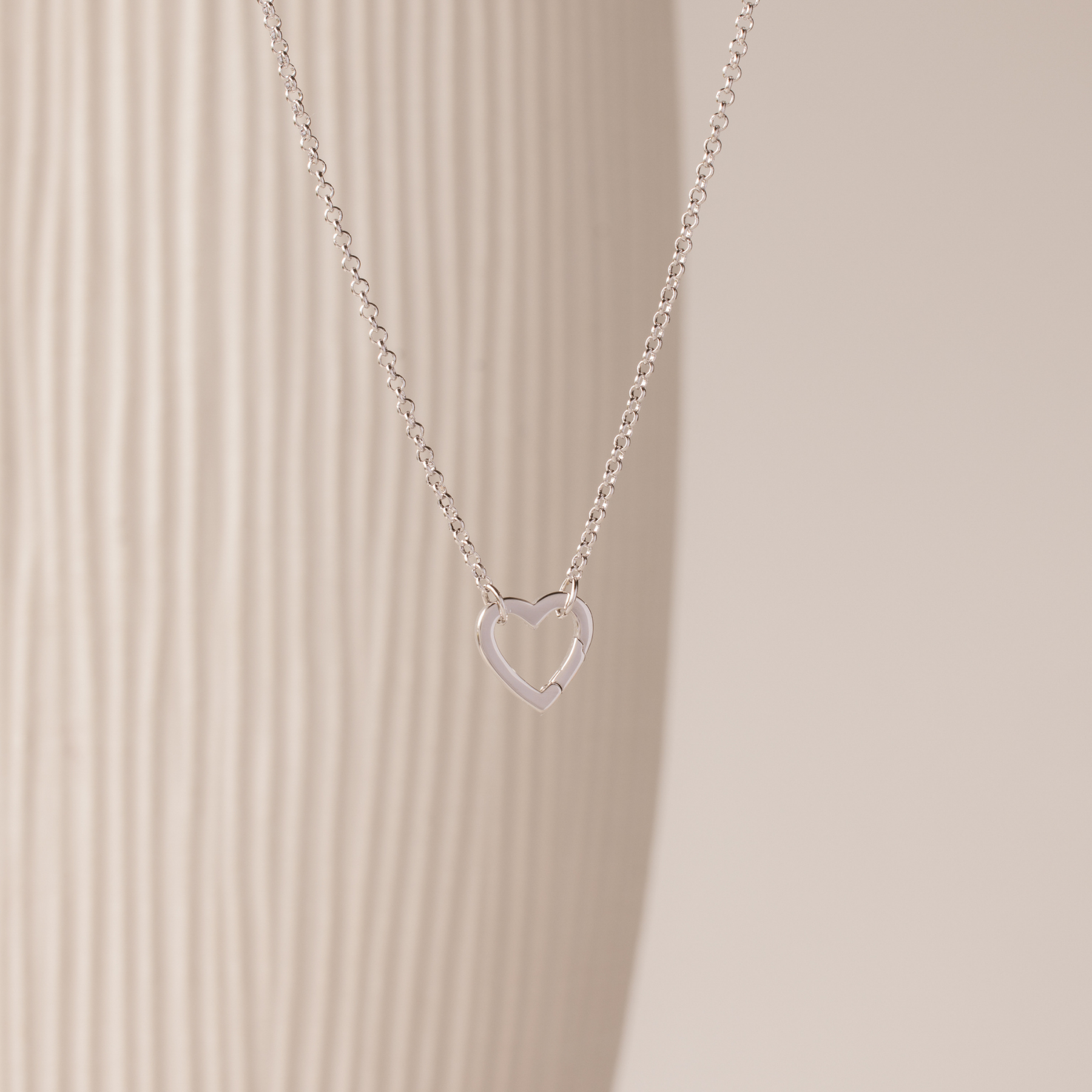 The Rolo Heart Clasp Necklace in White Gold features a delicate open heart pendant on a classic rolo chain, shown against a neutral textured background.