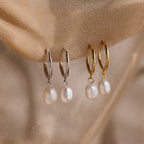Two pairs of gold and silver hoop earrings with pearl drops on a beige fabric background