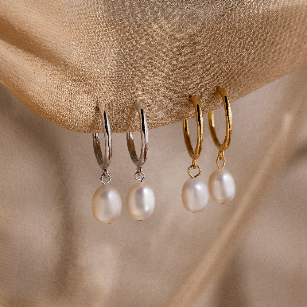 Two pairs of gold and silver hoop earrings with pearl drops on a beige fabric background