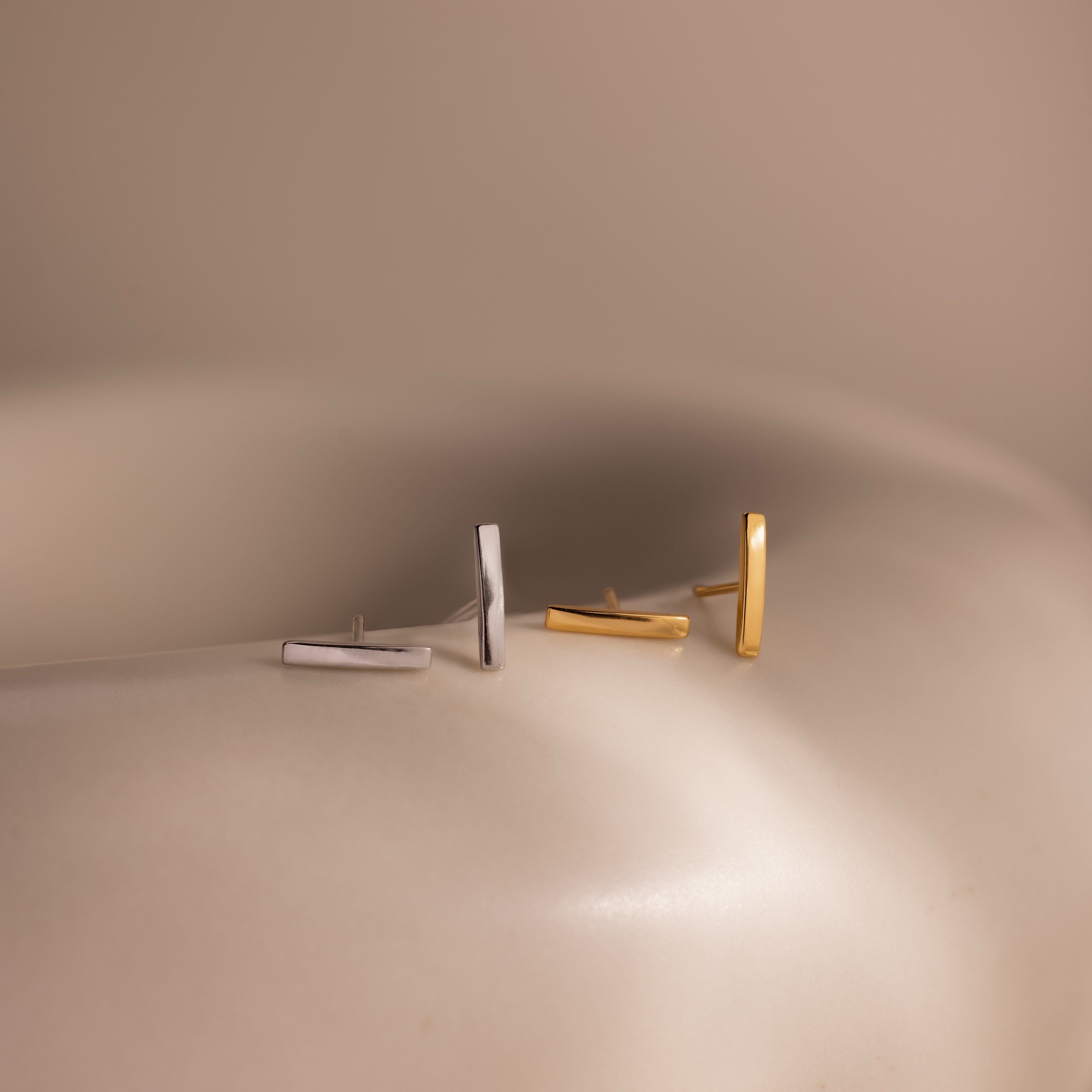 Two pairs of bar stud earrings, one silver and one gold, on a neutral background