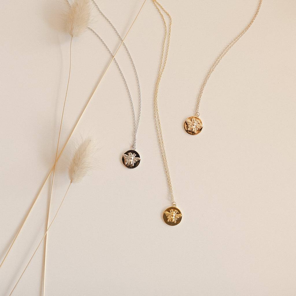 Three Bee Coin Necklaces—one gold, one silver, and one rose gold—are displayed on a beige surface with dried grass accents, making them the perfect nature-inspired layering necklaces.