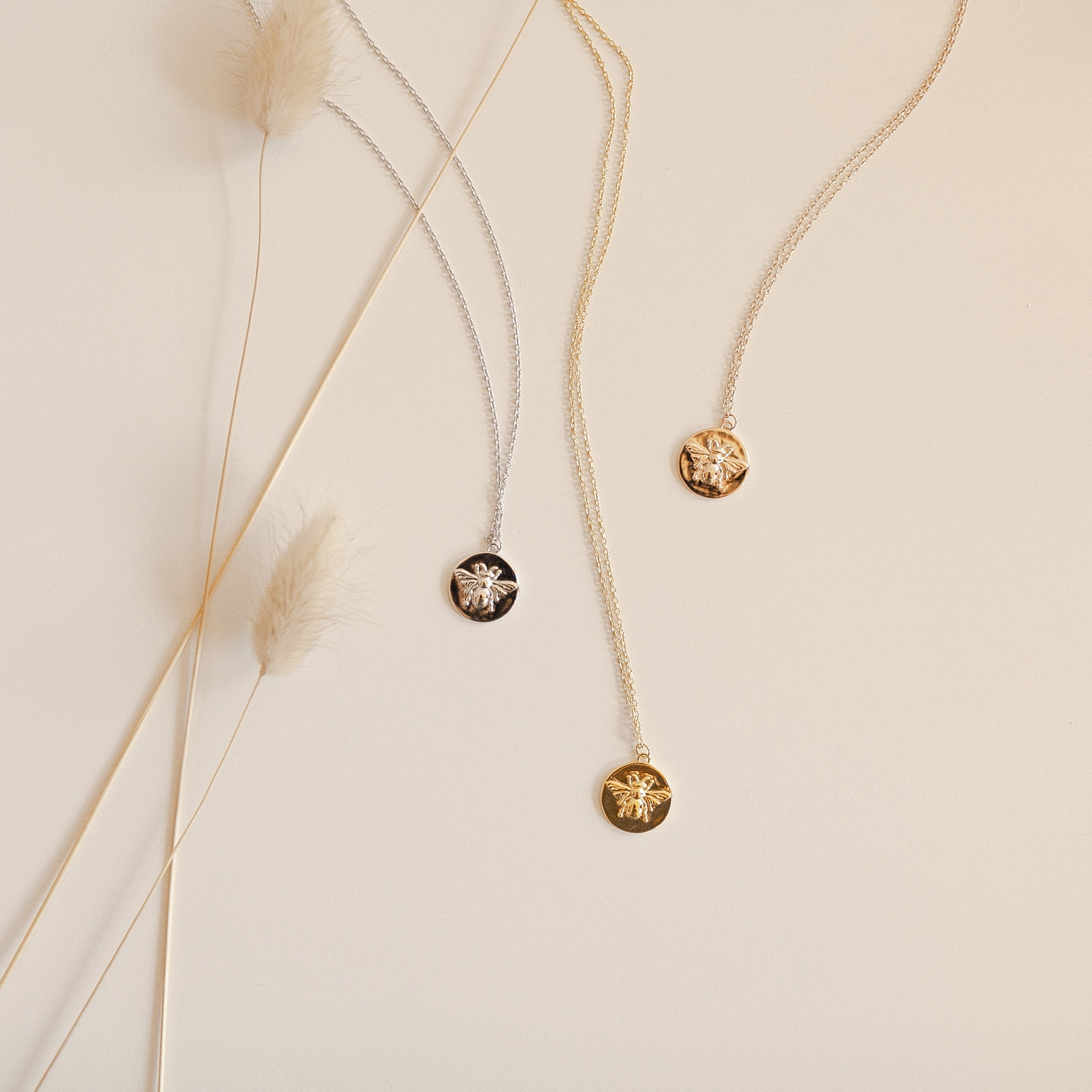 Three Bee Coin Necklaces—one gold, one silver, and one rose gold—are displayed on a beige surface with dried grass accents, making them the perfect nature-inspired layering necklaces.