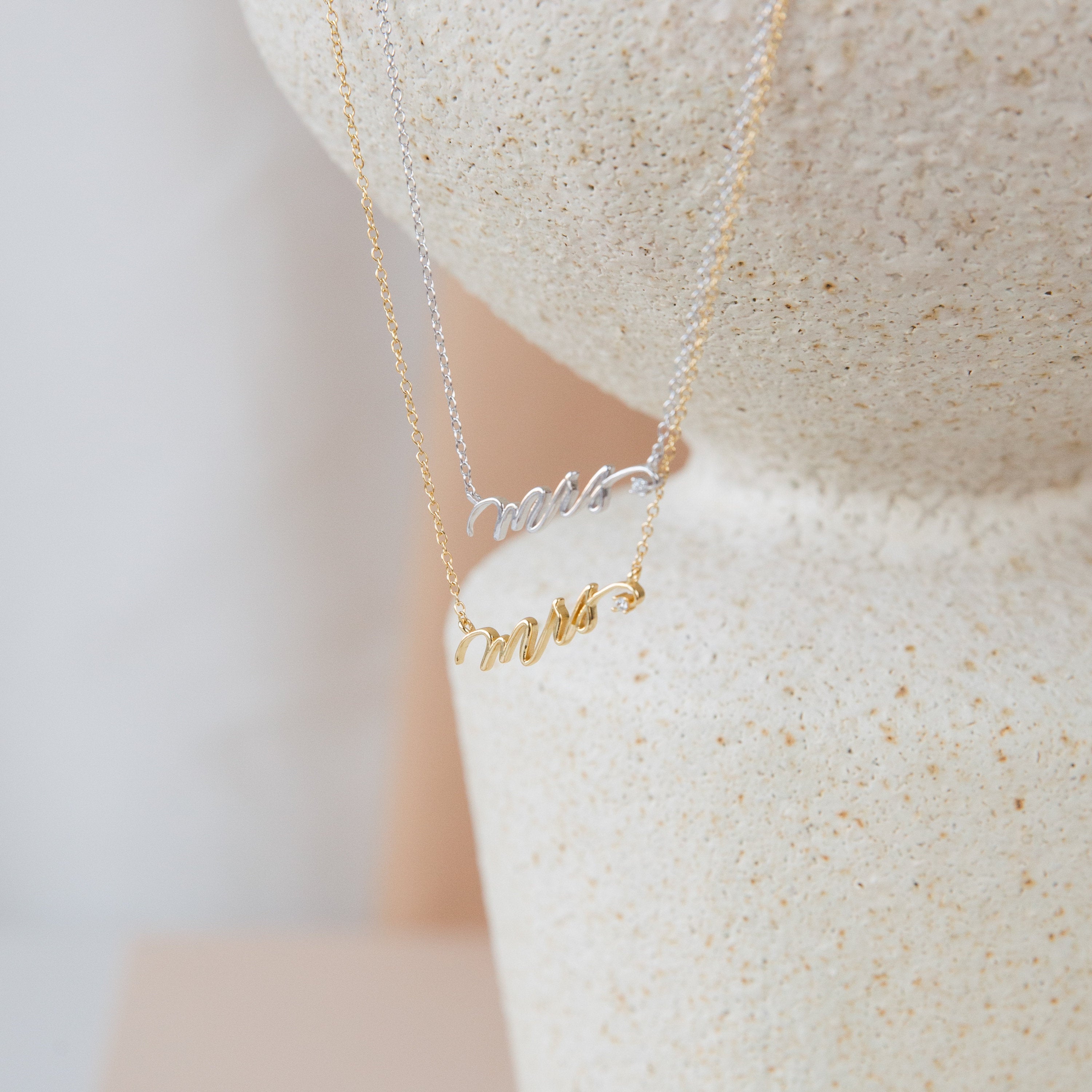 The Mrs. Necklace in silver and gold rests on a textured, light surface—an elegant wedding gift for any bride-to-be.