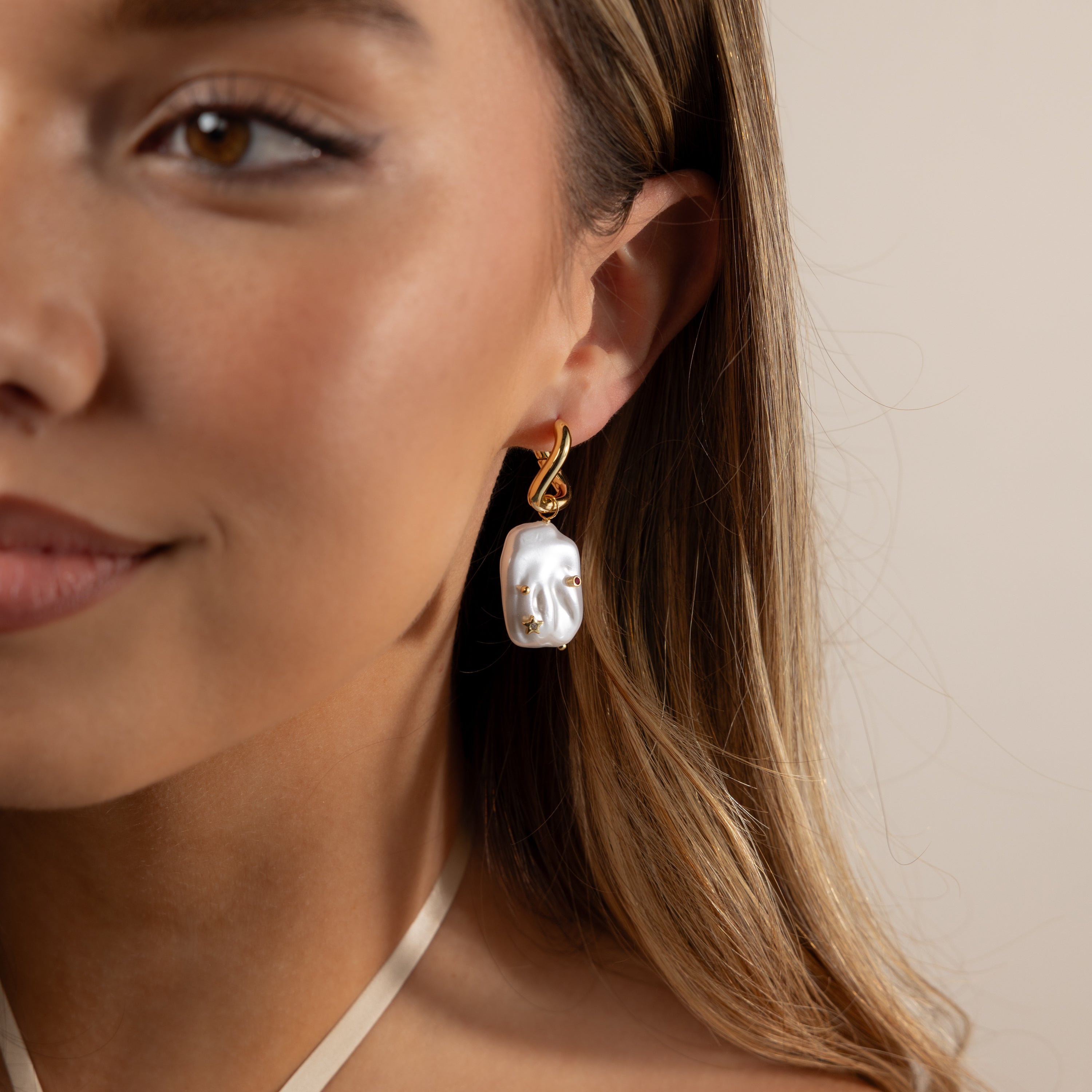A woman wears Celestial Baroque Pearl Charm Hoops in 18K Gold, featuring a large, irregular baroque pearl, shown close up on her face and ear.