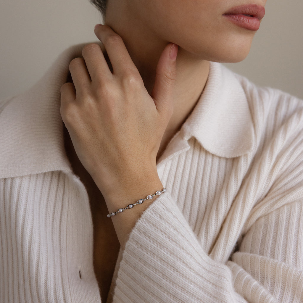 A person in a white sweater touches their neck, elegantly displaying the Eternity Pearl Bracelet in Sterling Silver alongside a delicate silver piece adorned with small stones.