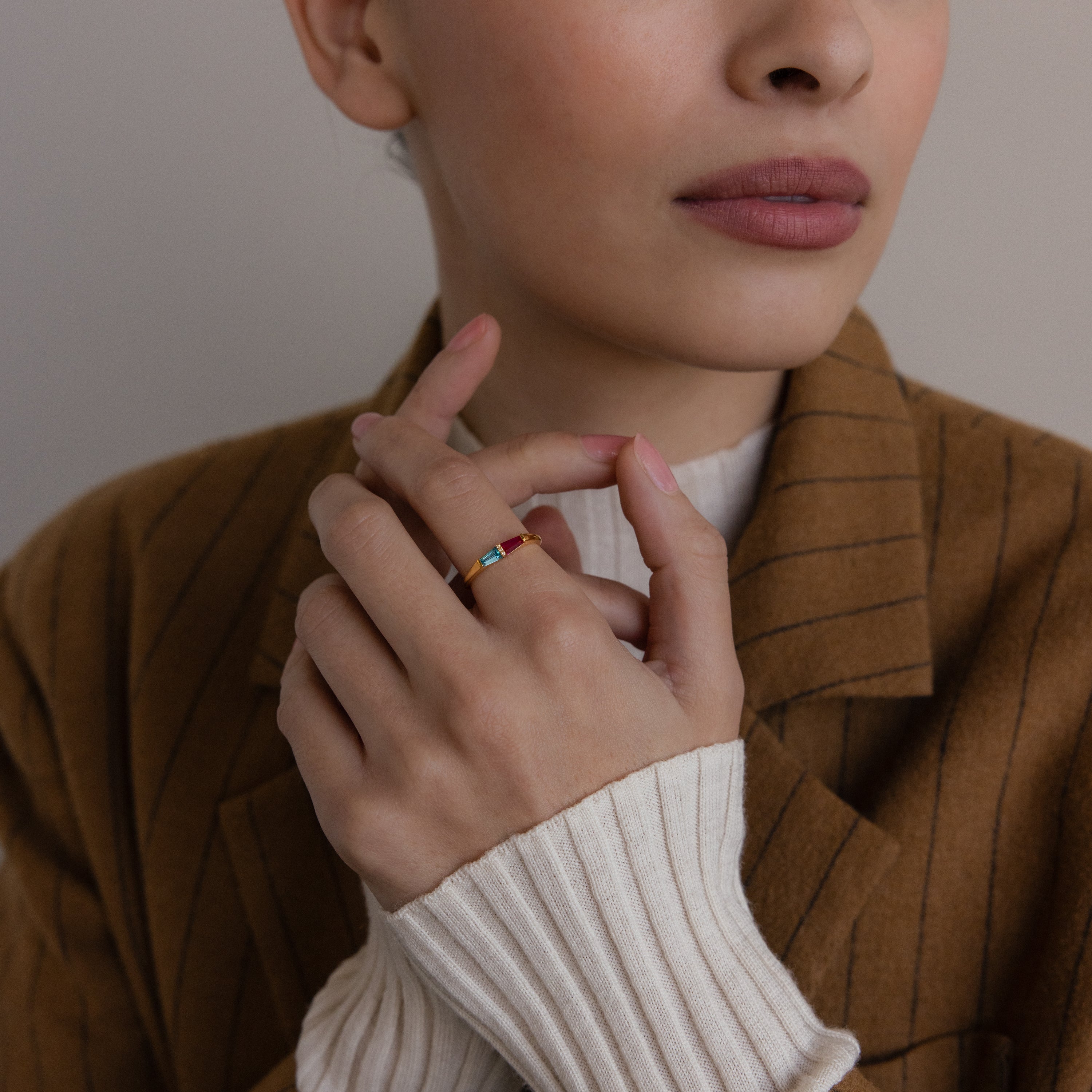 A woman in a brown pinstripe blazer touches her face, showcasing the vibrant Toi et Moi Tapered Birthstone Ring on her hand.
