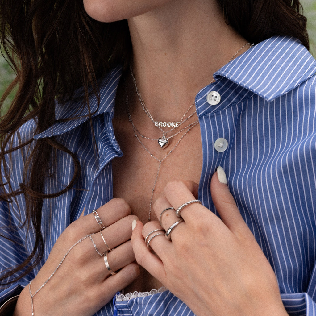Woman wearing a blue pinstripe shirt, layered silver necklaces including the Tiny Bubble Name Necklace in Silver, and multiple rings on her fingers.