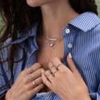 Woman in a blue striped shirt wears layered rings and the Tiny Bubble Name Necklace, holding her collar with manicured hands.