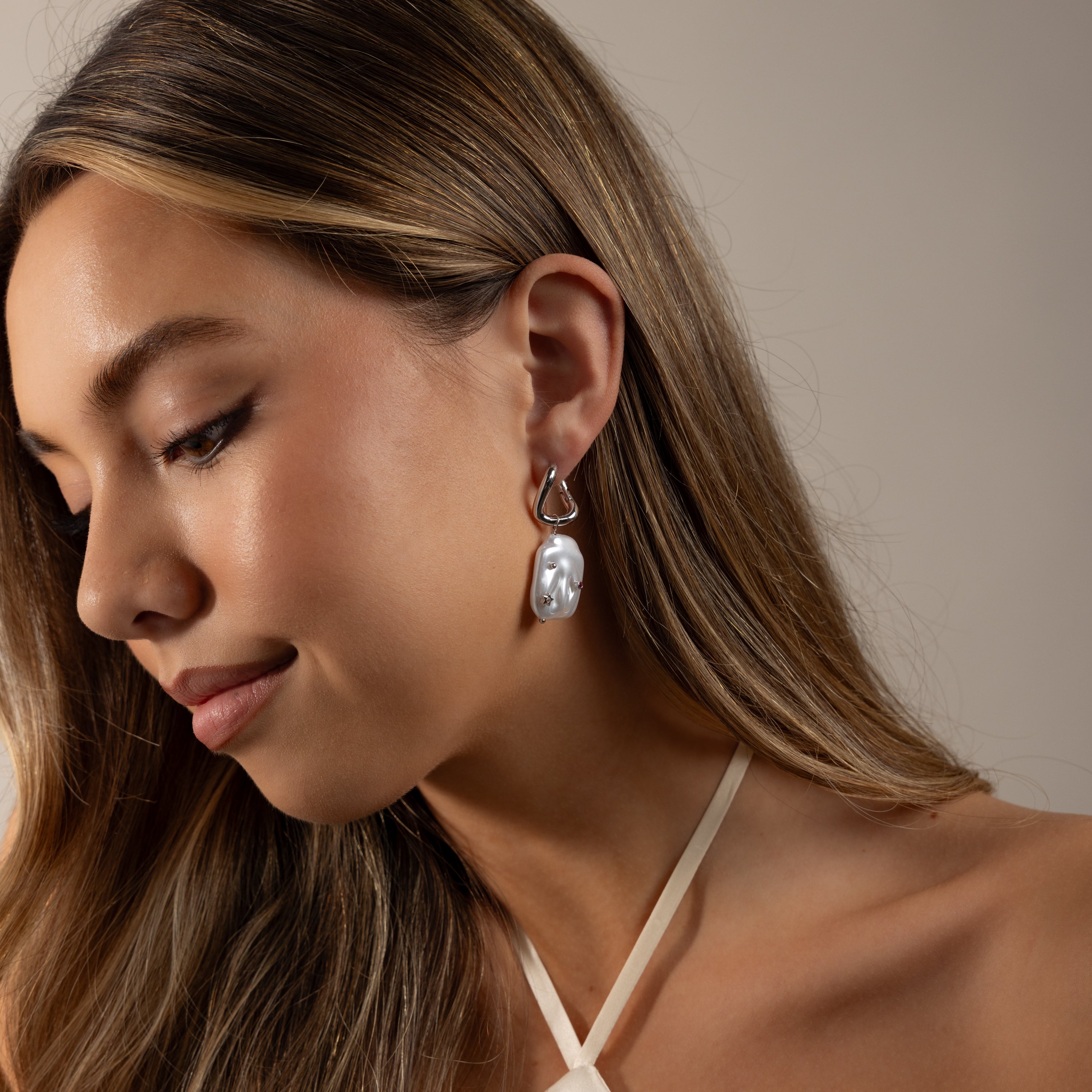 Woman with long hair smiles softly, looking down while wearing Celestial Baroque Pearl Charm Hoops in White Gold and a halter top.