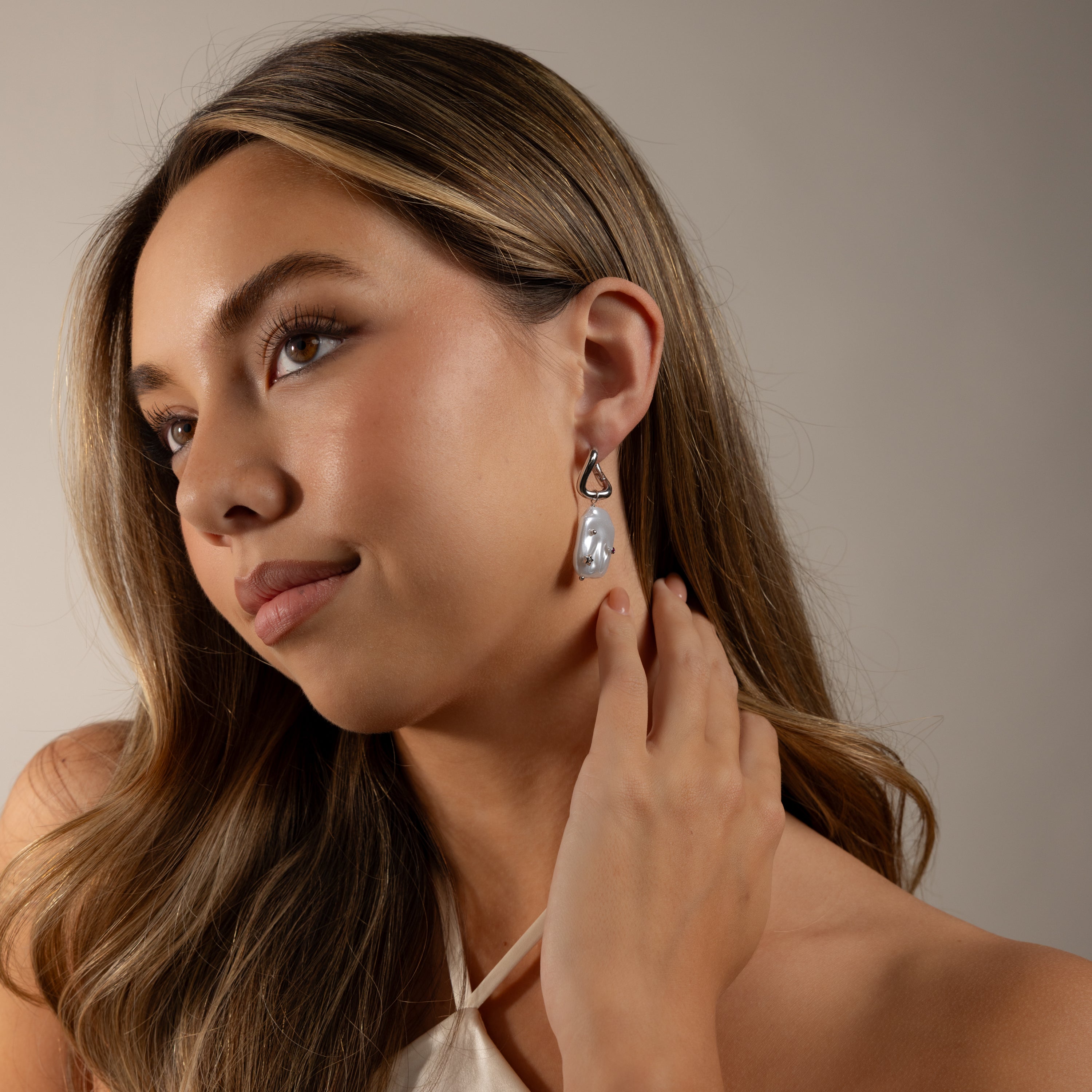 Woman with long hair in a white top holds the Celestial Baroque Pearl Charm Hoops in White Gold, gazing slightly upward.
