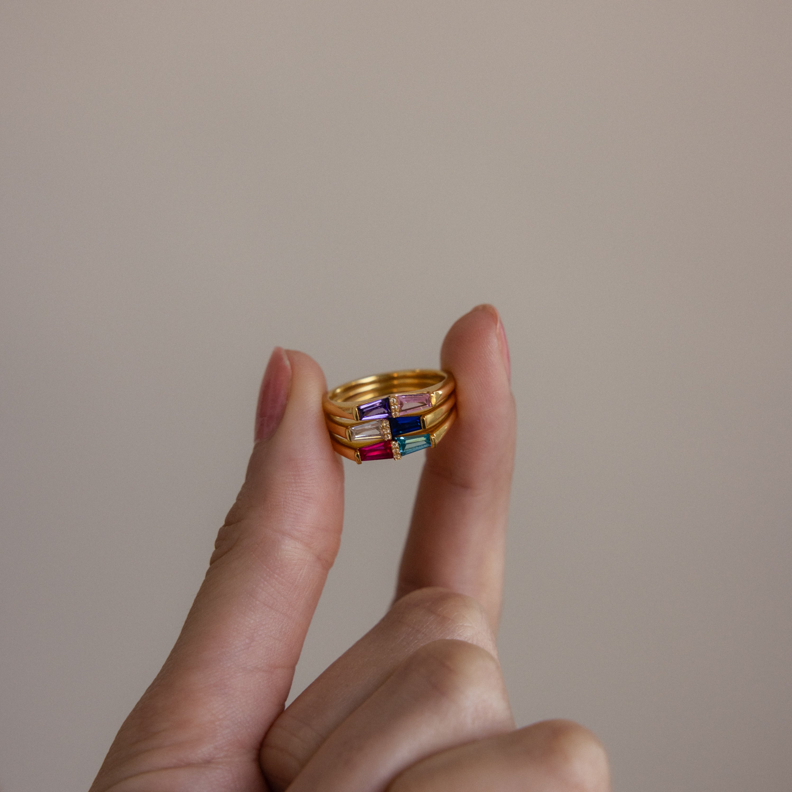 A hand displays three stacked Toi et Moi Tapered Birthstone Rings with vibrant gemstones, including a baguette-cut birthstone, set against a plain beige background.