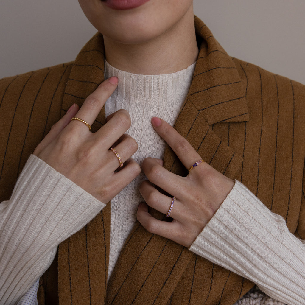 A woman wearing gold rings, including the Toi et Moi Tapered Birthstone Ring, poses with her hands on her chest while dressed in a brown pinstripe blazer and cream sweater.