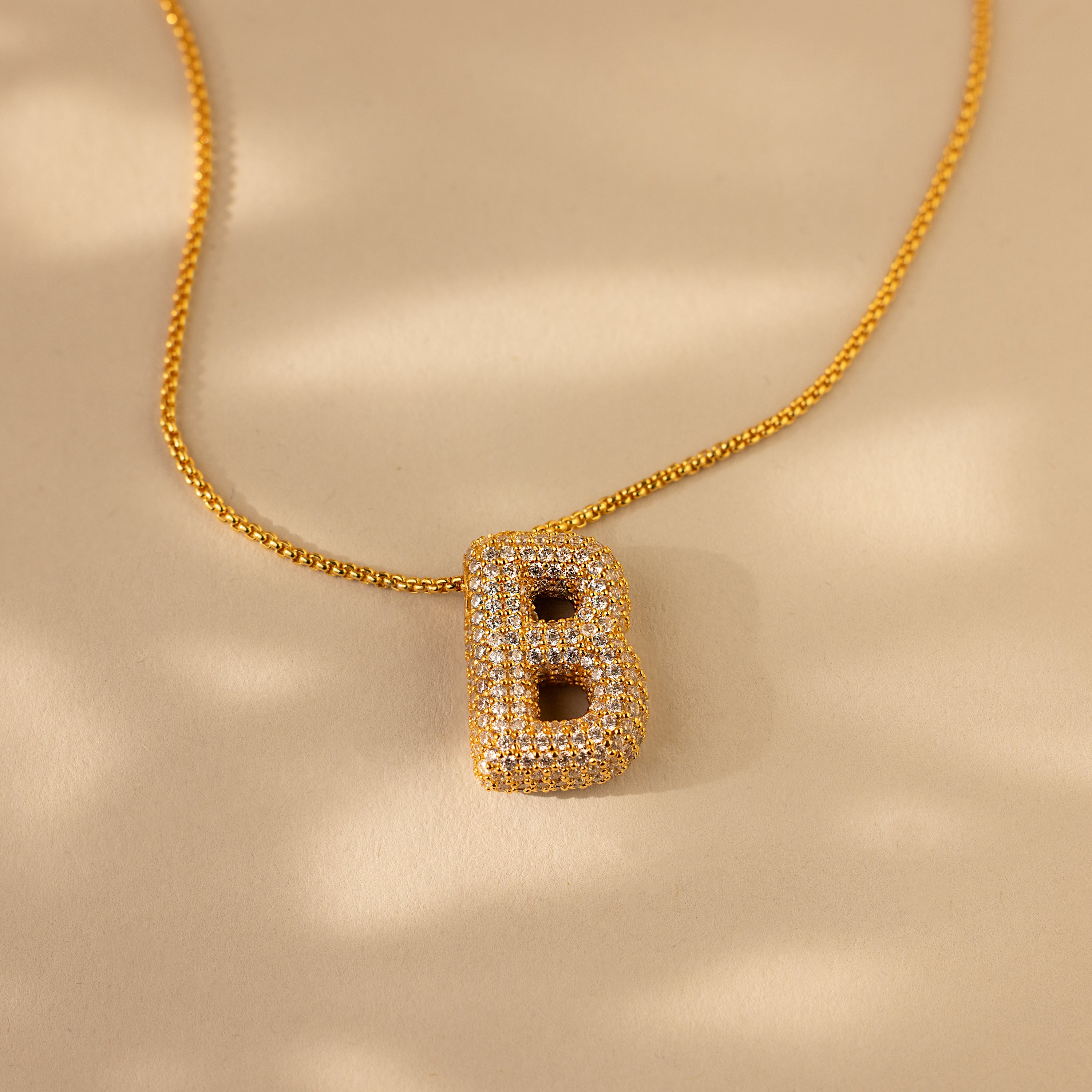 Pave Puff Letter Necklace in 18K Gold featuring a diamond-studded letter B pendant, displayed on a beige background with soft shadows.