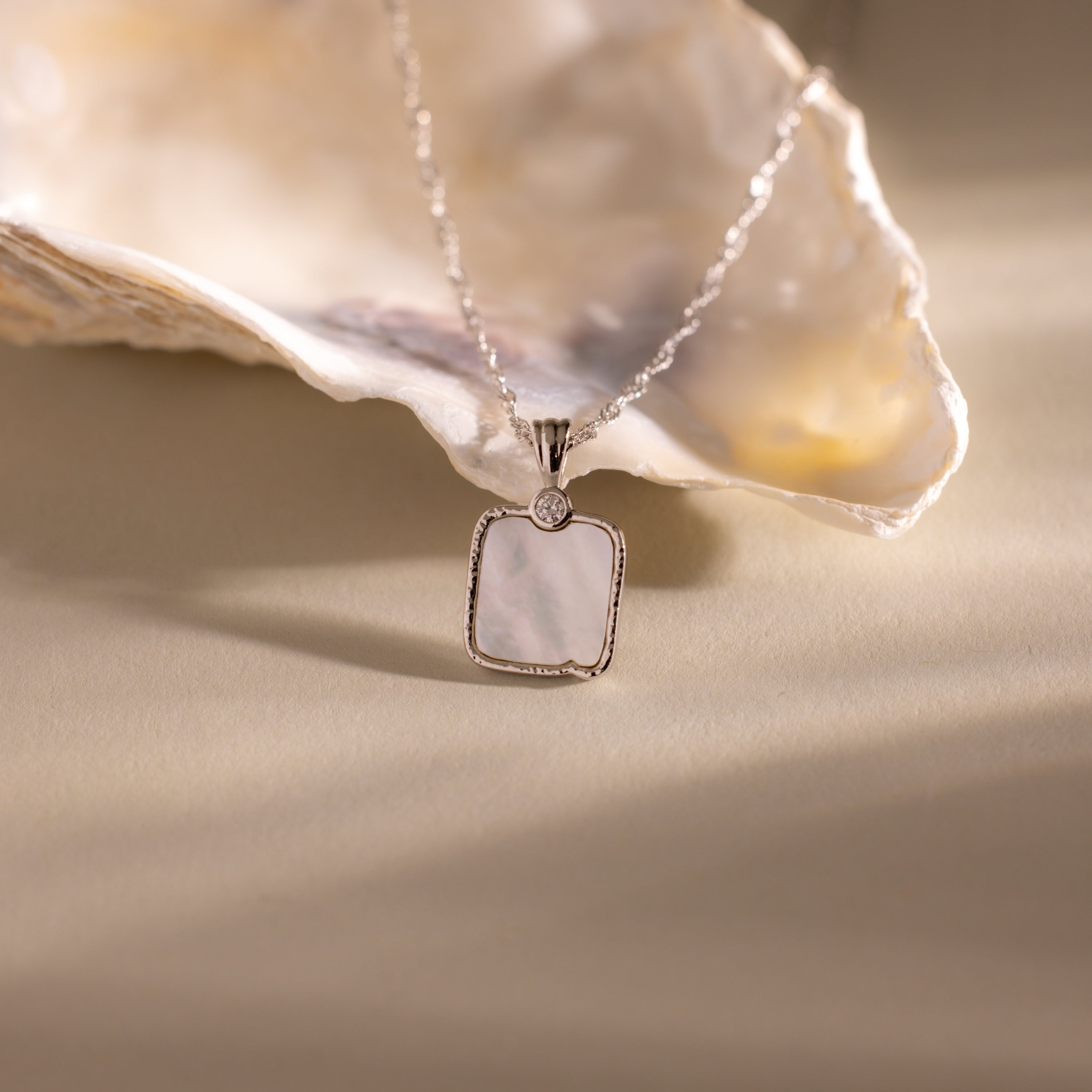 The Square Mother of Pearl Necklace features an iridescent pendant and is displayed before an open seashell, highlighting its elegant and delicate design.