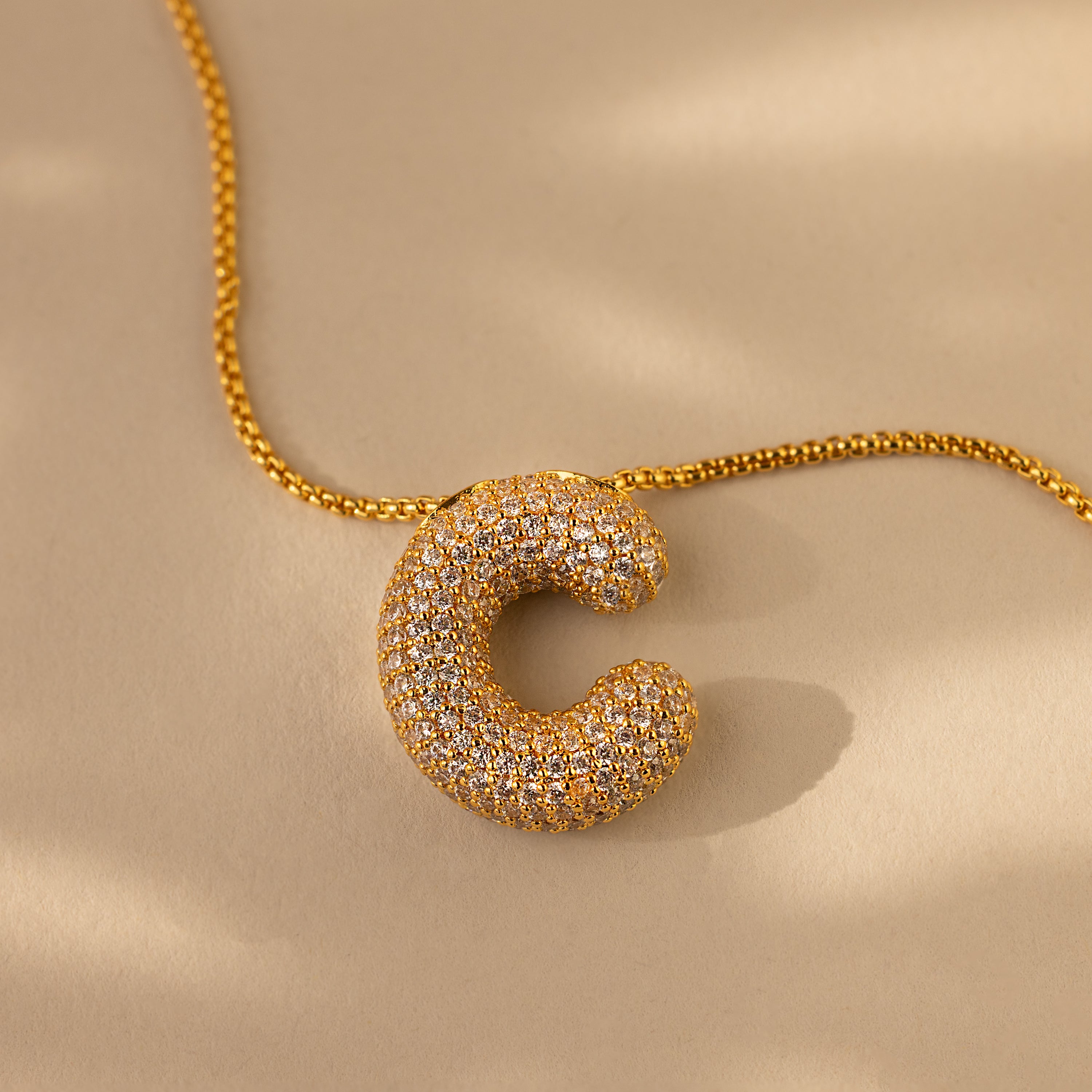 The Pave Puff Letter Necklace in 18K Gold features a C-shaped pendant encrusted with sparkling pavé diamonds, displayed on a neutral background.