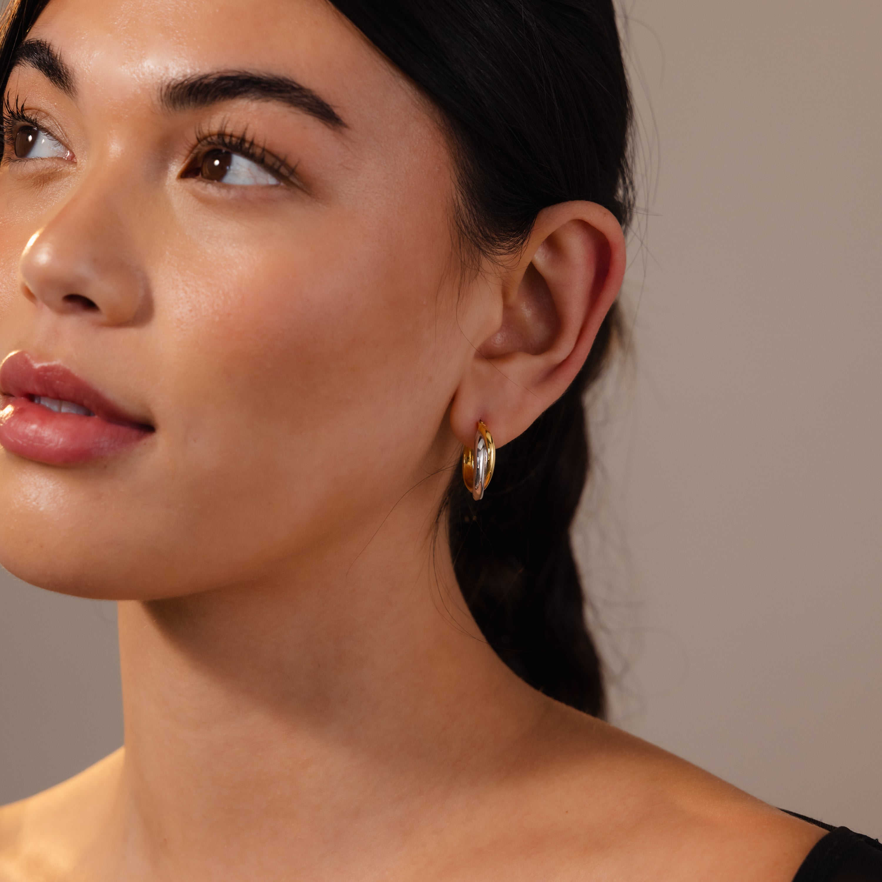 Woman with smooth skin wearing Twisted Mixed Metal Hoops, looking upwards against a neutral background.
