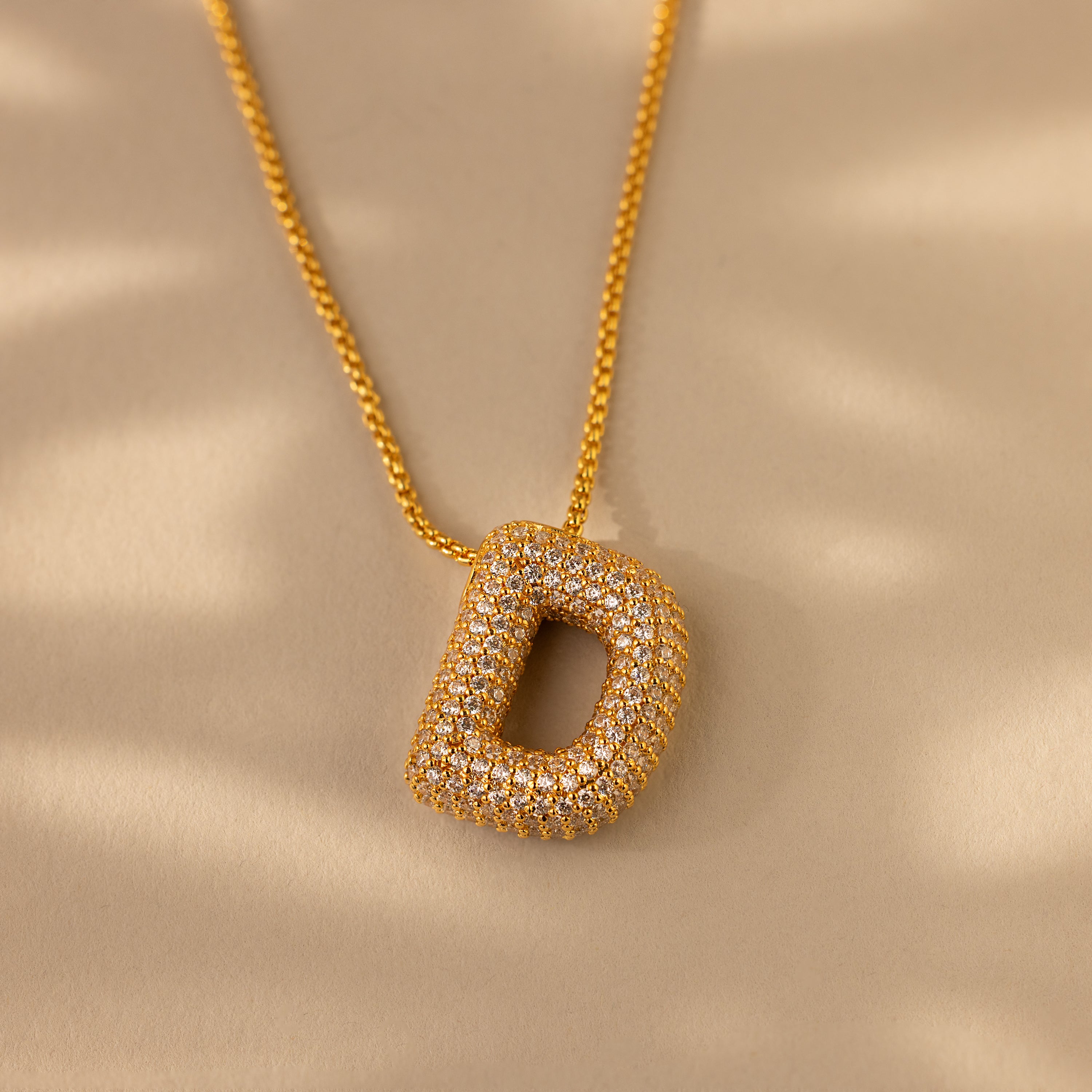 The Pave Puff Letter Necklace in 18K Gold showcases a sparkling, pave diamonds-studded letter D pendant, beautifully displayed on a beige background.