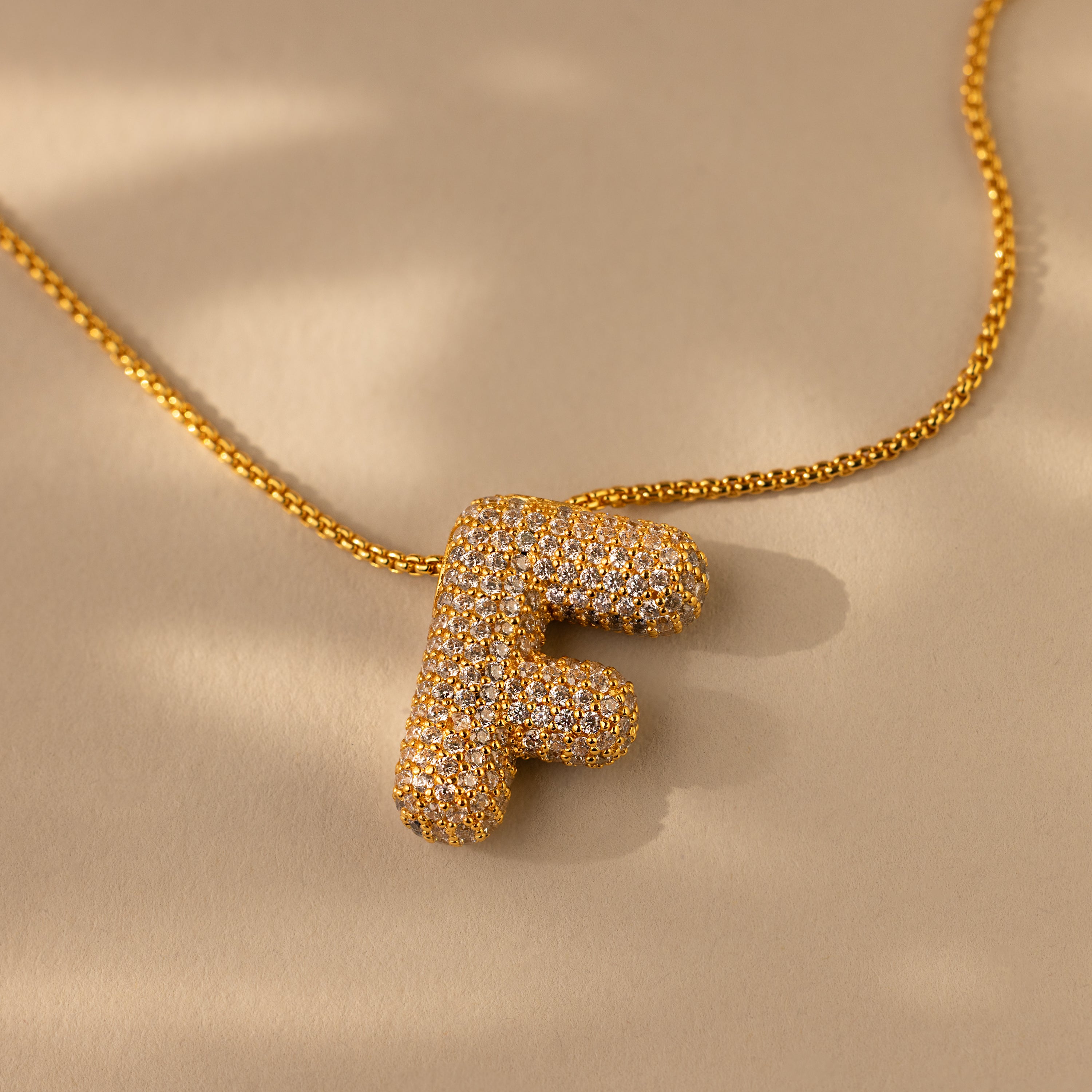 The Pave Puff Letter Necklace in 18K Gold features a pendant shaped like the letter F with pave diamonds, shown on a beige background.