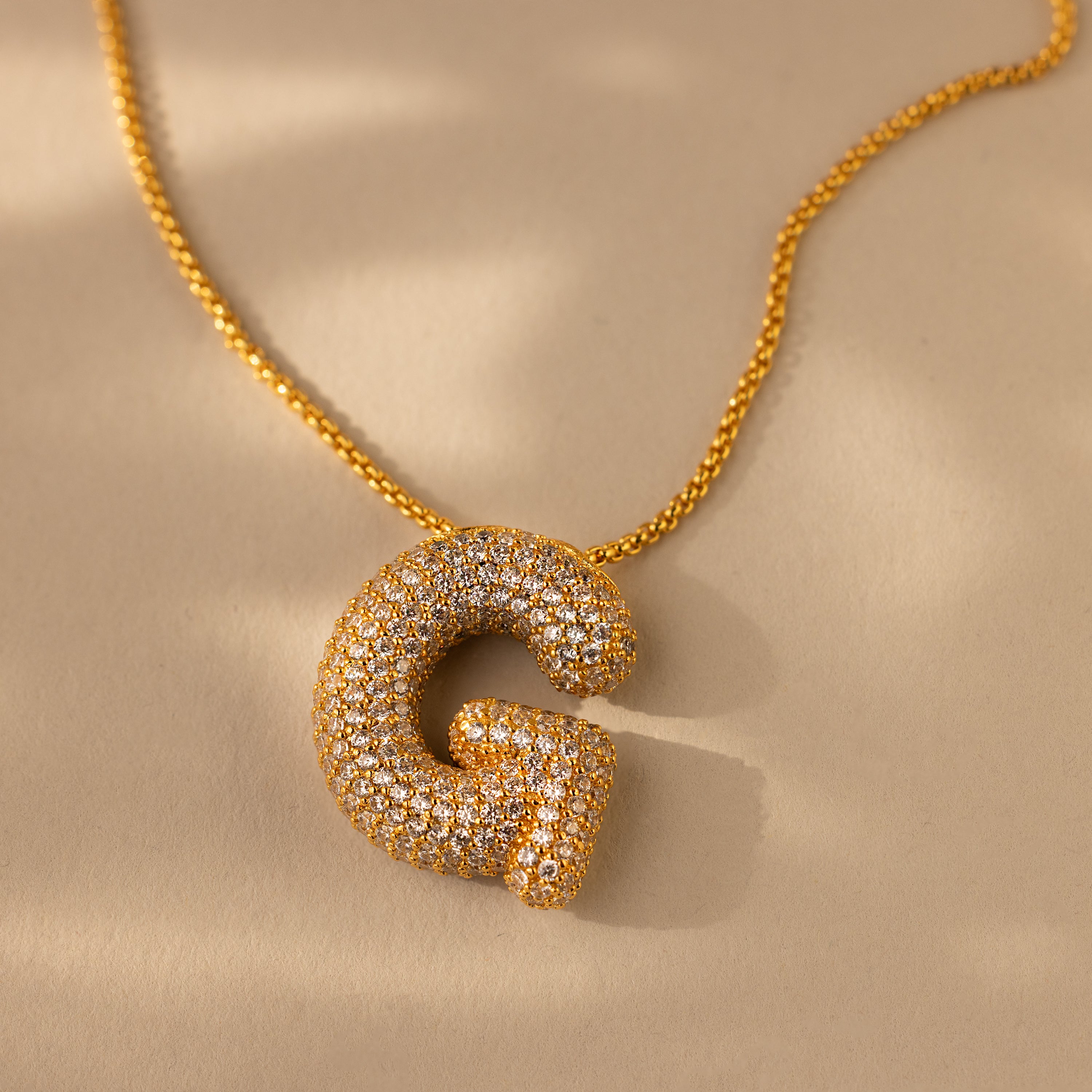 The Pave Puff Letter Necklace in 18K Gold features a diamond-encrusted letter G pendant, showcased on a beige background.