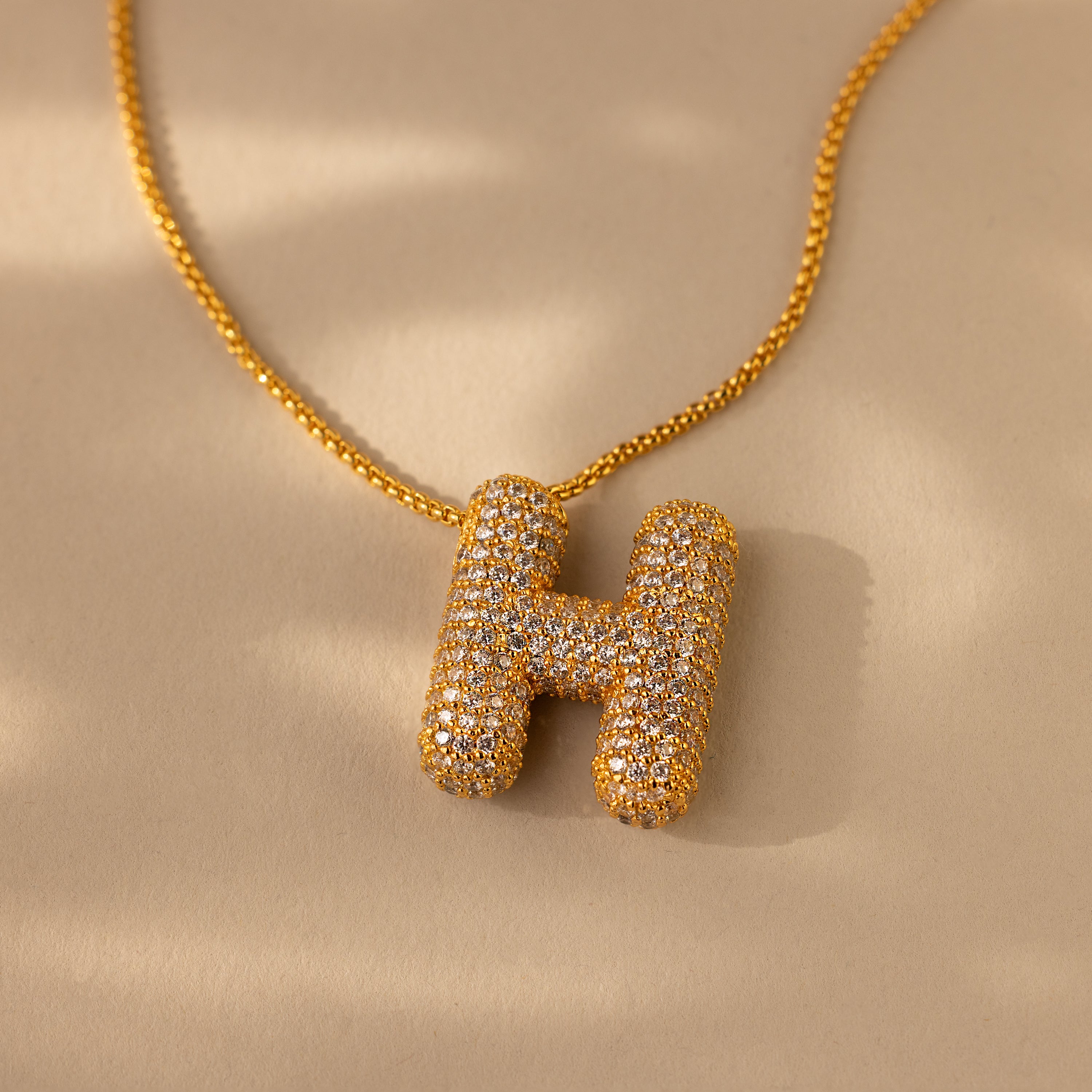 The Pave Puff Letter Necklace in 18K Gold showcases a sparkling letter H pendant with pave diamonds, set against a beige background.