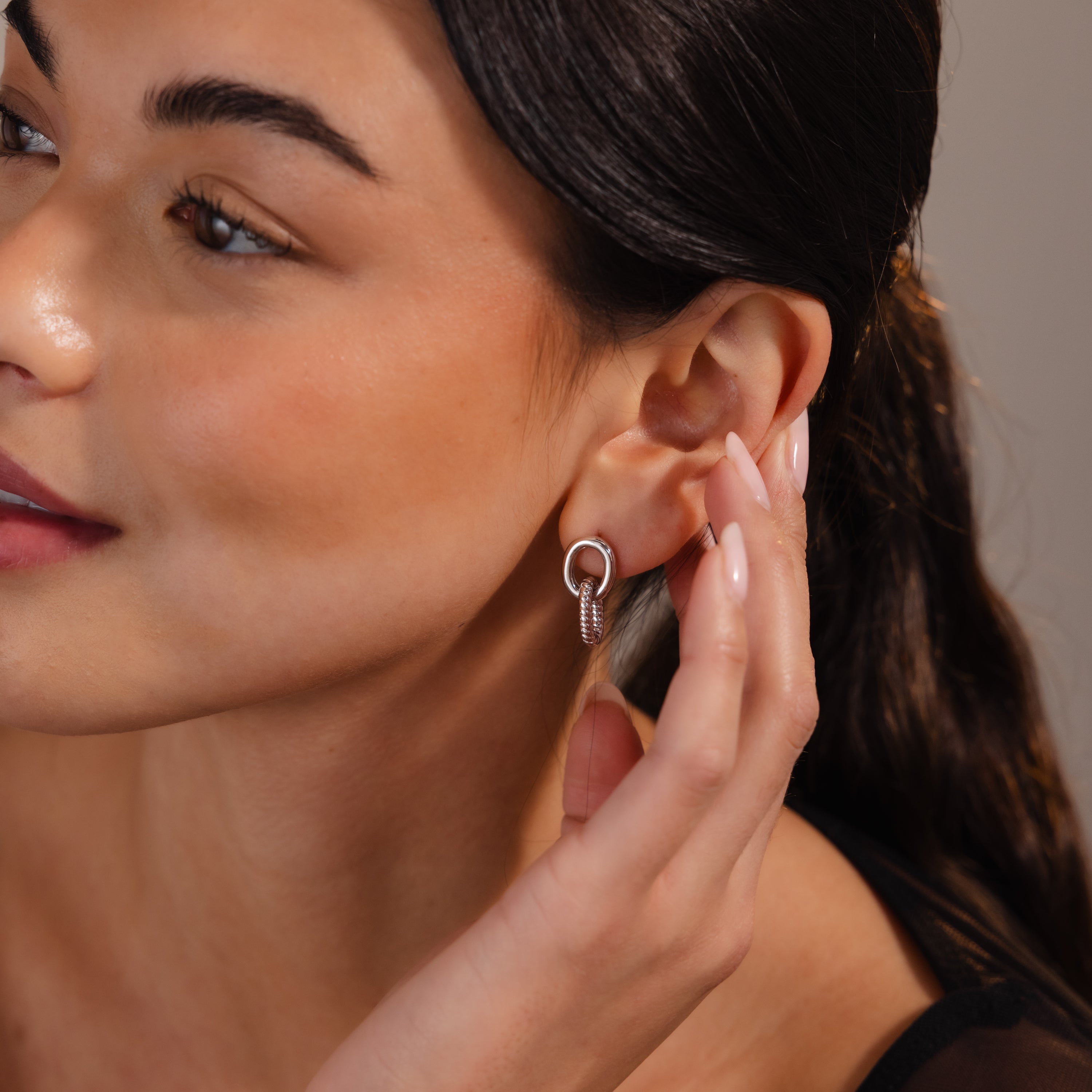 A woman touches her earring, highlighting the Twisted Link Earrings in White Gold—a modern yet timeless jewelry piece featuring elegant silver hoops with a unique knot design.