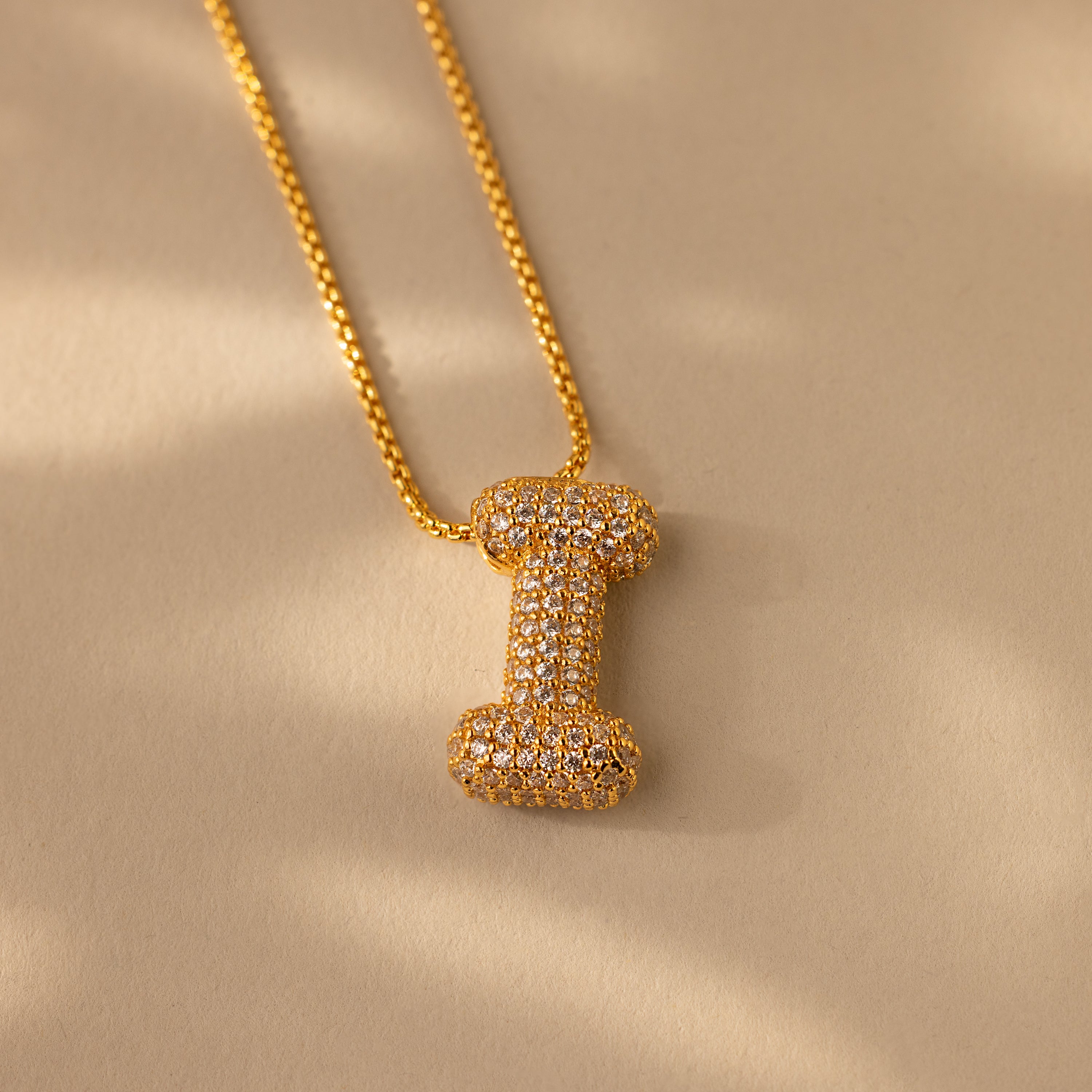 The Pave Puff Letter Necklace in 18K Gold features a diamond-studded “I” charm, displayed against a beige background.
