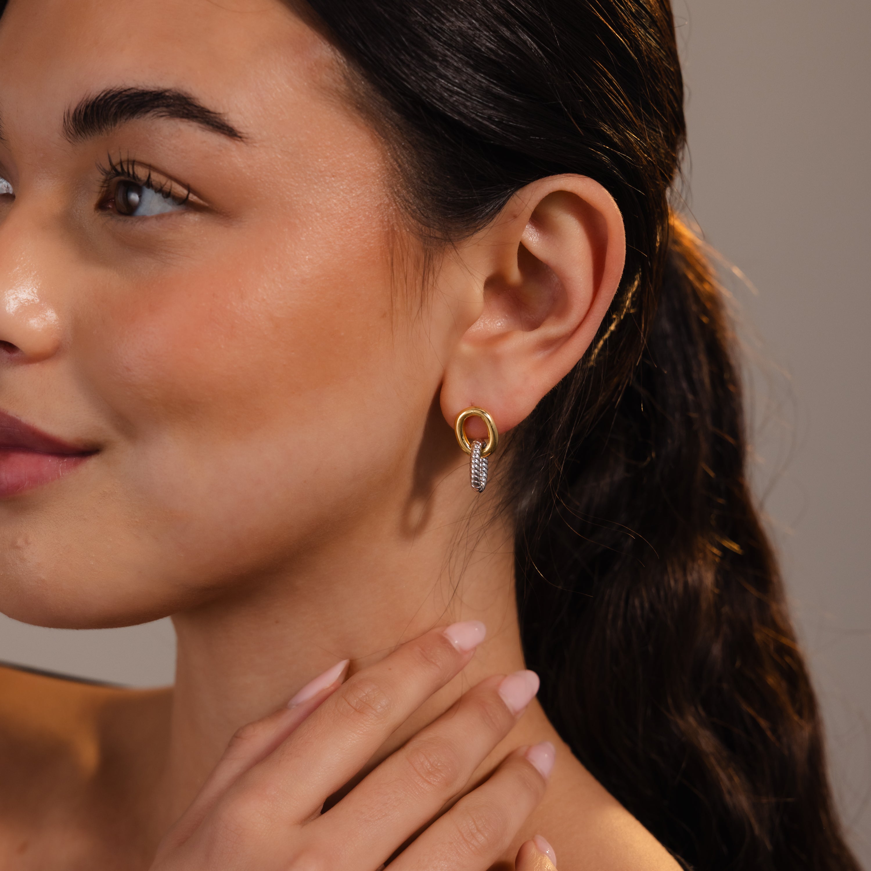 A woman with long dark hair gently touches her neck while wearing sculptural gold Twisted Link Earrings, which feature a decorative pendant.