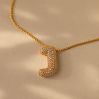 The Pave Puff Letter Necklace in 18K Gold showcases a sparkling pave diamond "J" charm on a beige background.