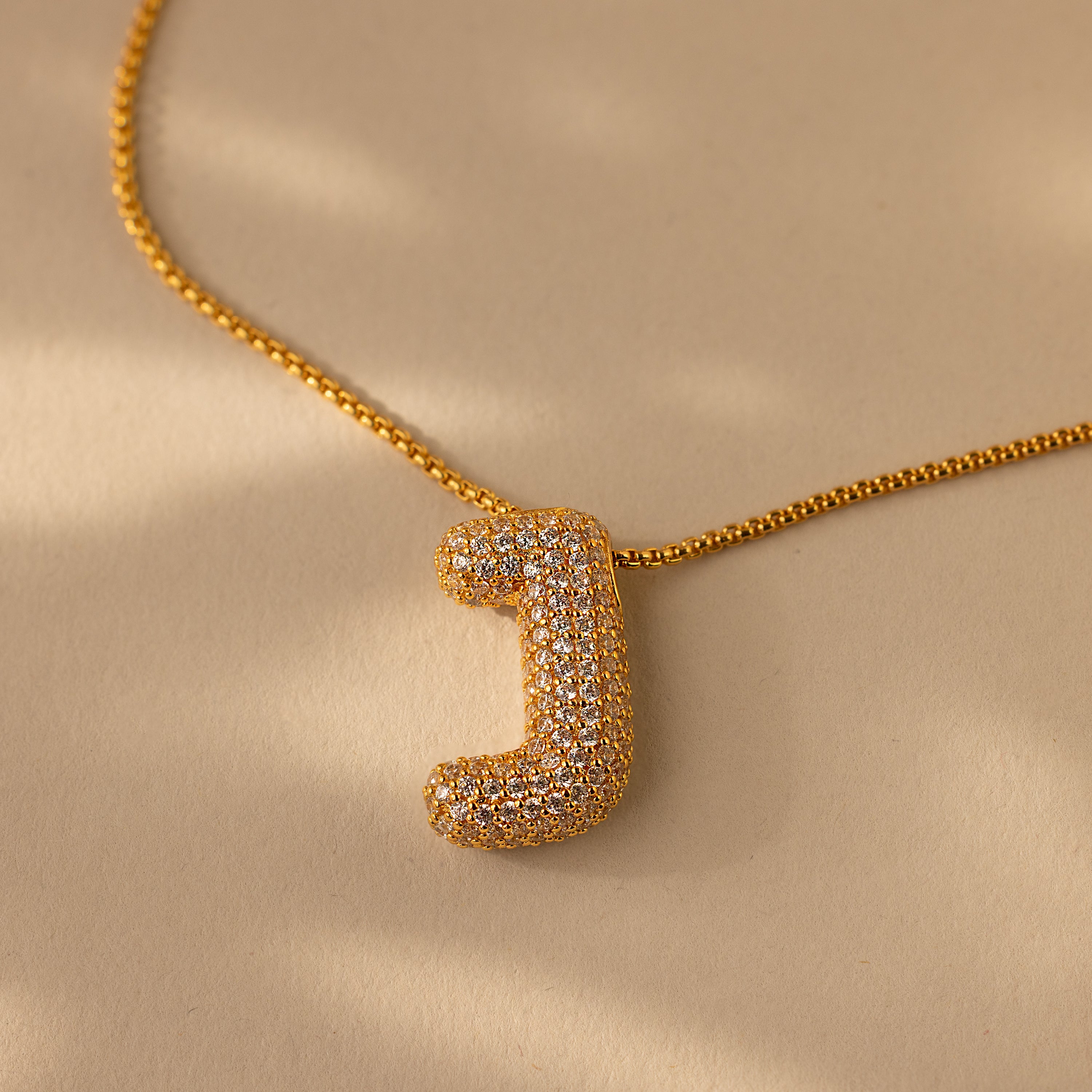 The Pave Puff Letter Necklace in 18K Gold showcases a sparkling pave diamond "J" charm on a beige background.