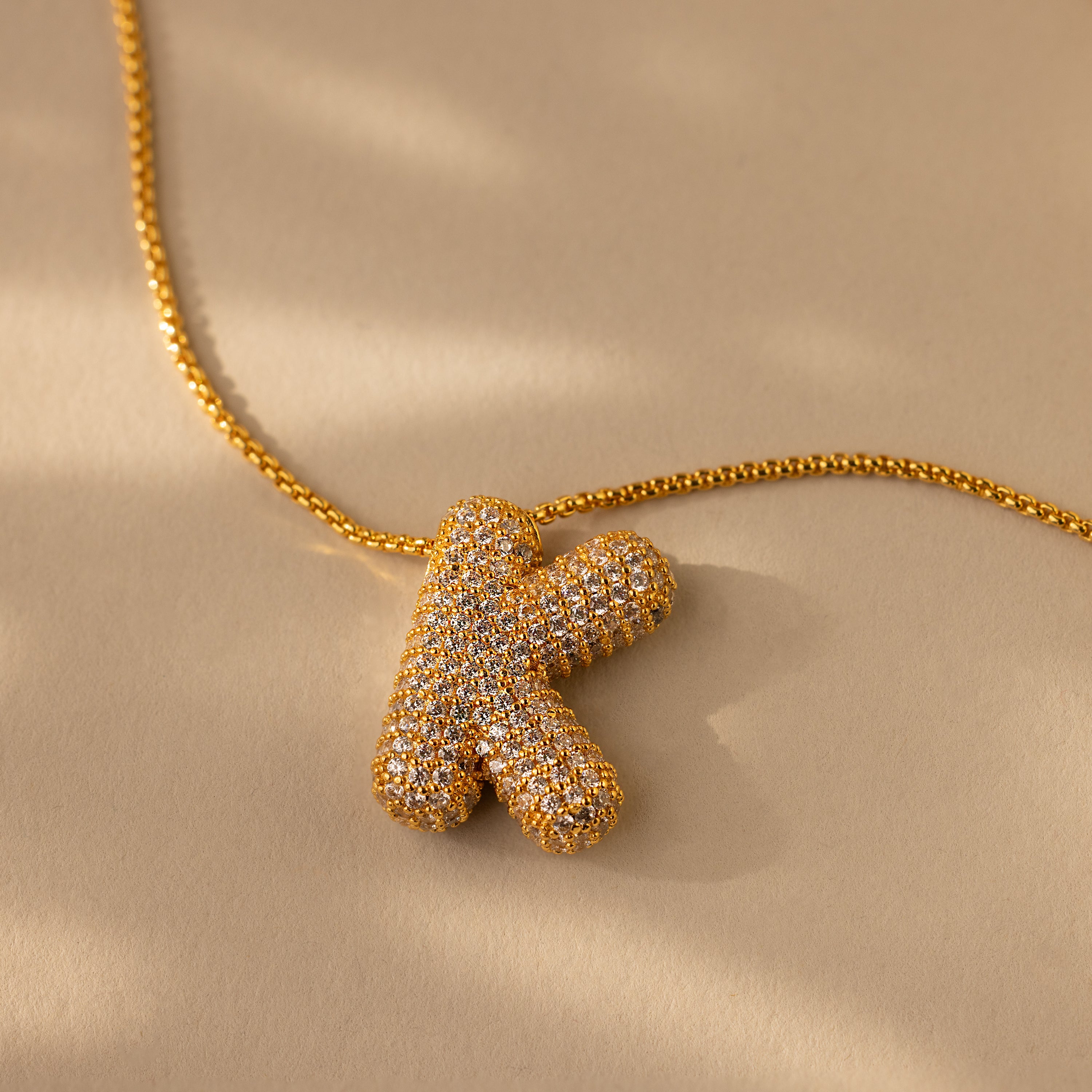 The Pave Puff Letter Necklace in 18K Gold showcases a gold "K" letter charm encrusted with pave diamonds, displayed on a beige background.