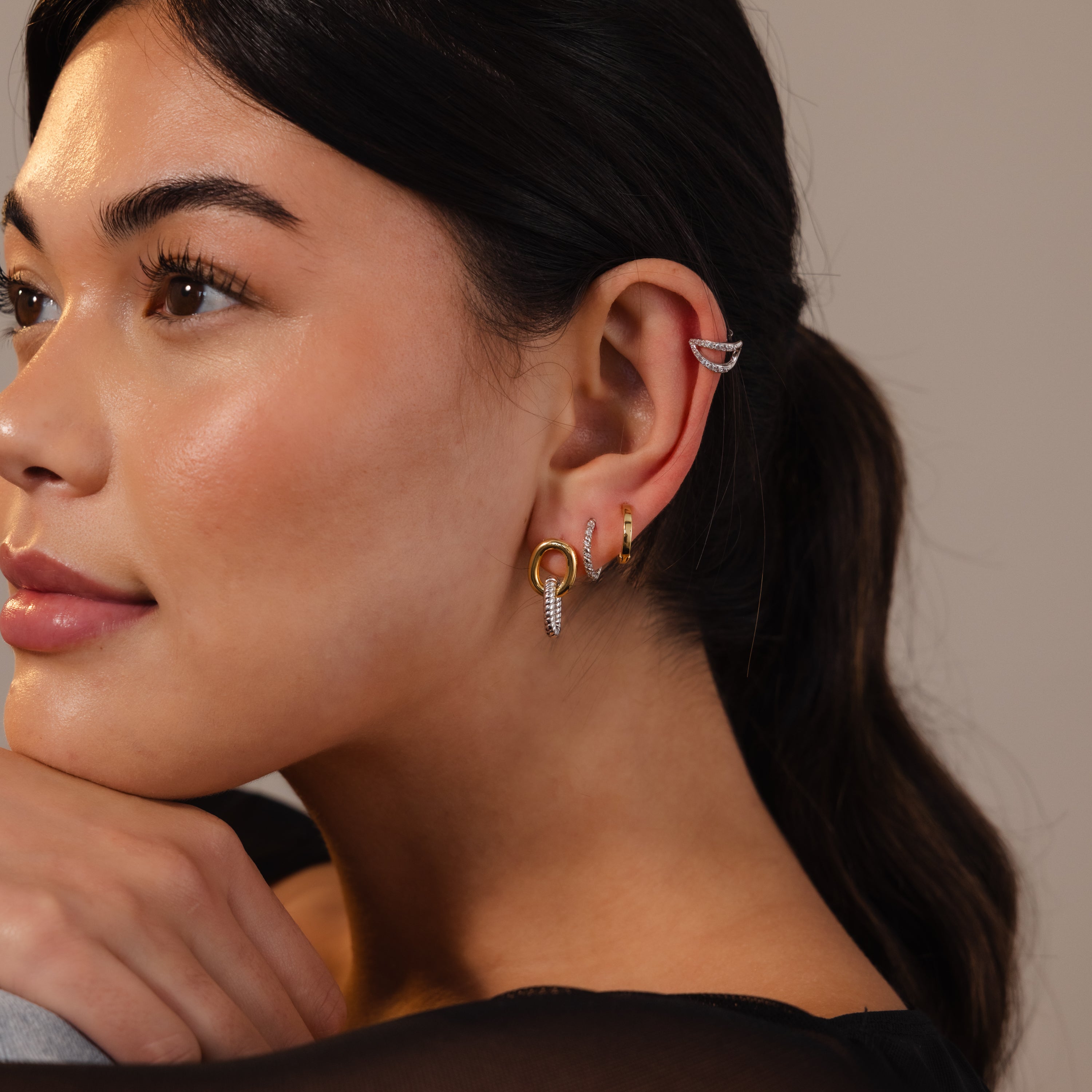 A woman with long dark hair wears multiple sculptural earrings, including the Twisted Link Earrings, as she gazes softly to the left.