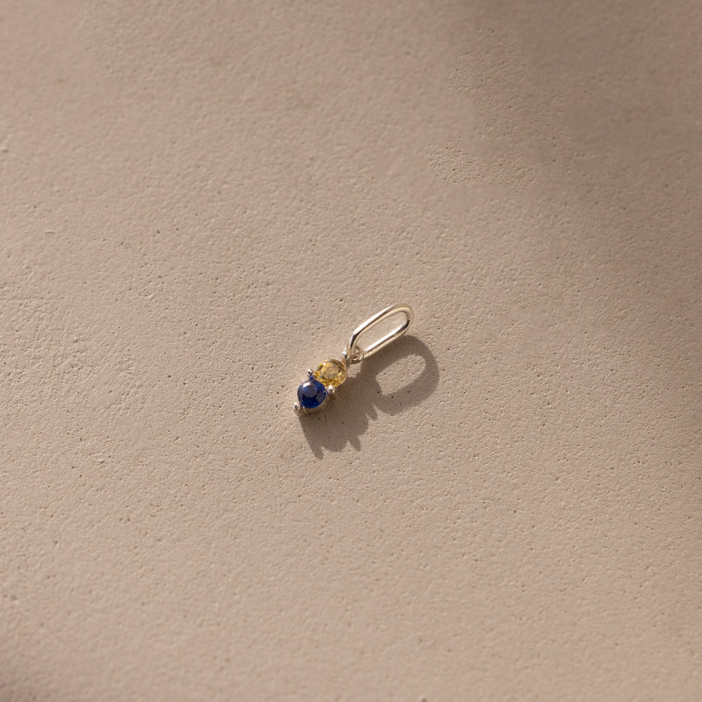 Silver Dainty Duo Birthstone Charm with topaz and sapphire. The design features two birthstones stacked vertically in prong setting with a large jump ring attached to the top. 