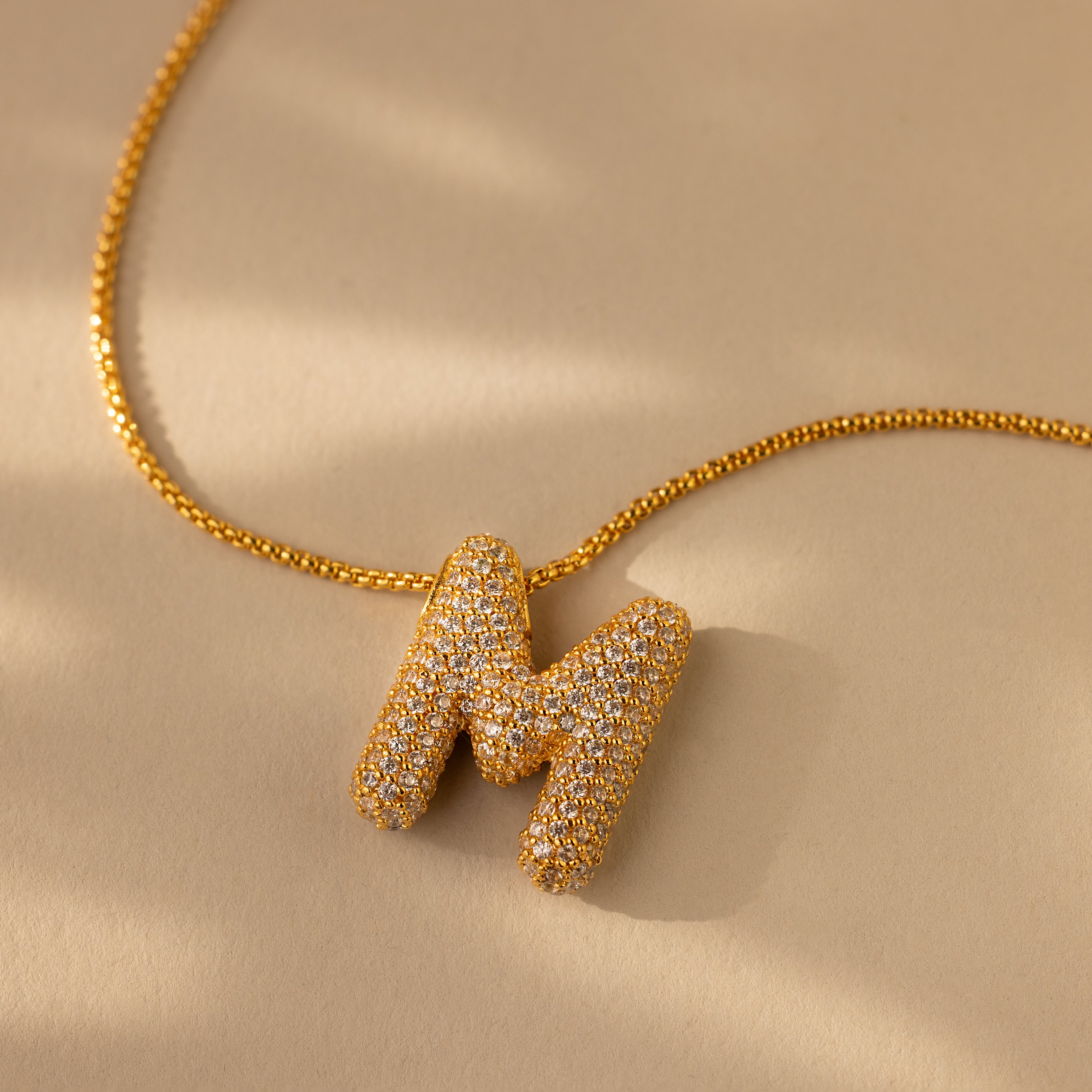 The Pave Puff Letter Necklace in 18K Gold features a sparkling, diamond-studded letter M pendant that shines with exquisite pave diamonds against a beige background.