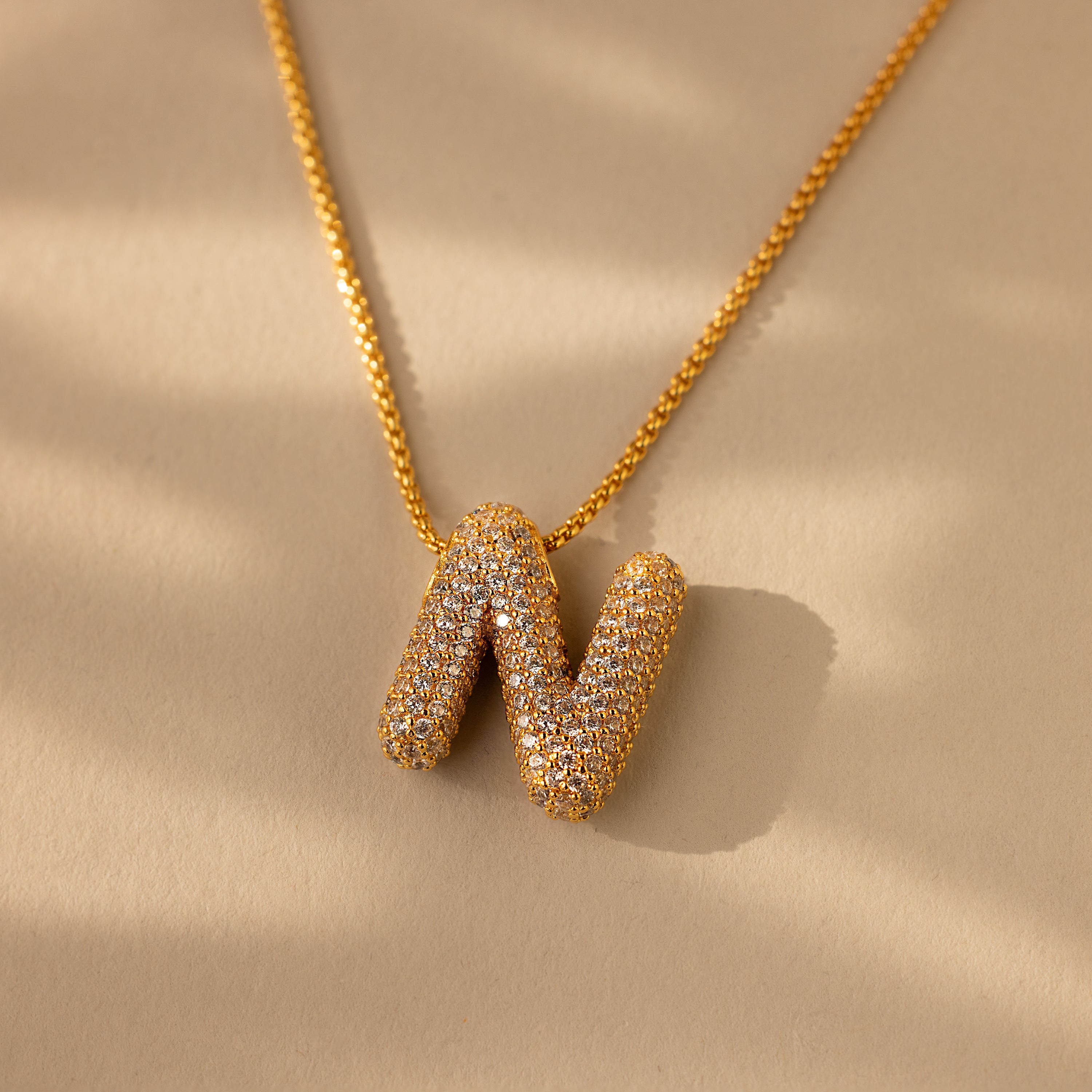 The Pave Puff Letter Necklace in 18K Gold showcases a gold letter N pendant with pave diamonds, set against a beige background.