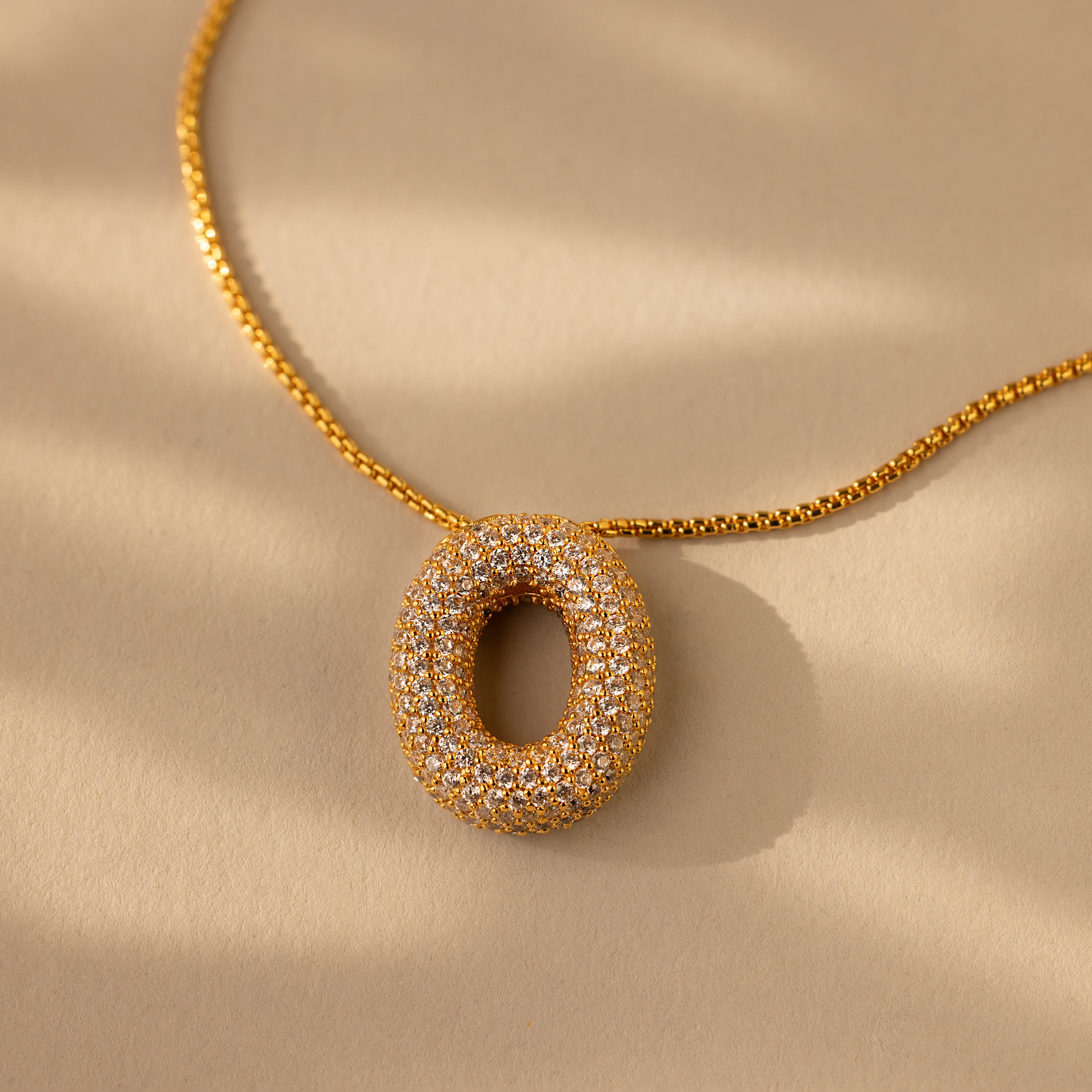 The Pave Puff Letter Necklace in 18K Gold features a circular, diamond-studded pendant set against a beige background with soft shadows, highlighting its pavé diamonds.