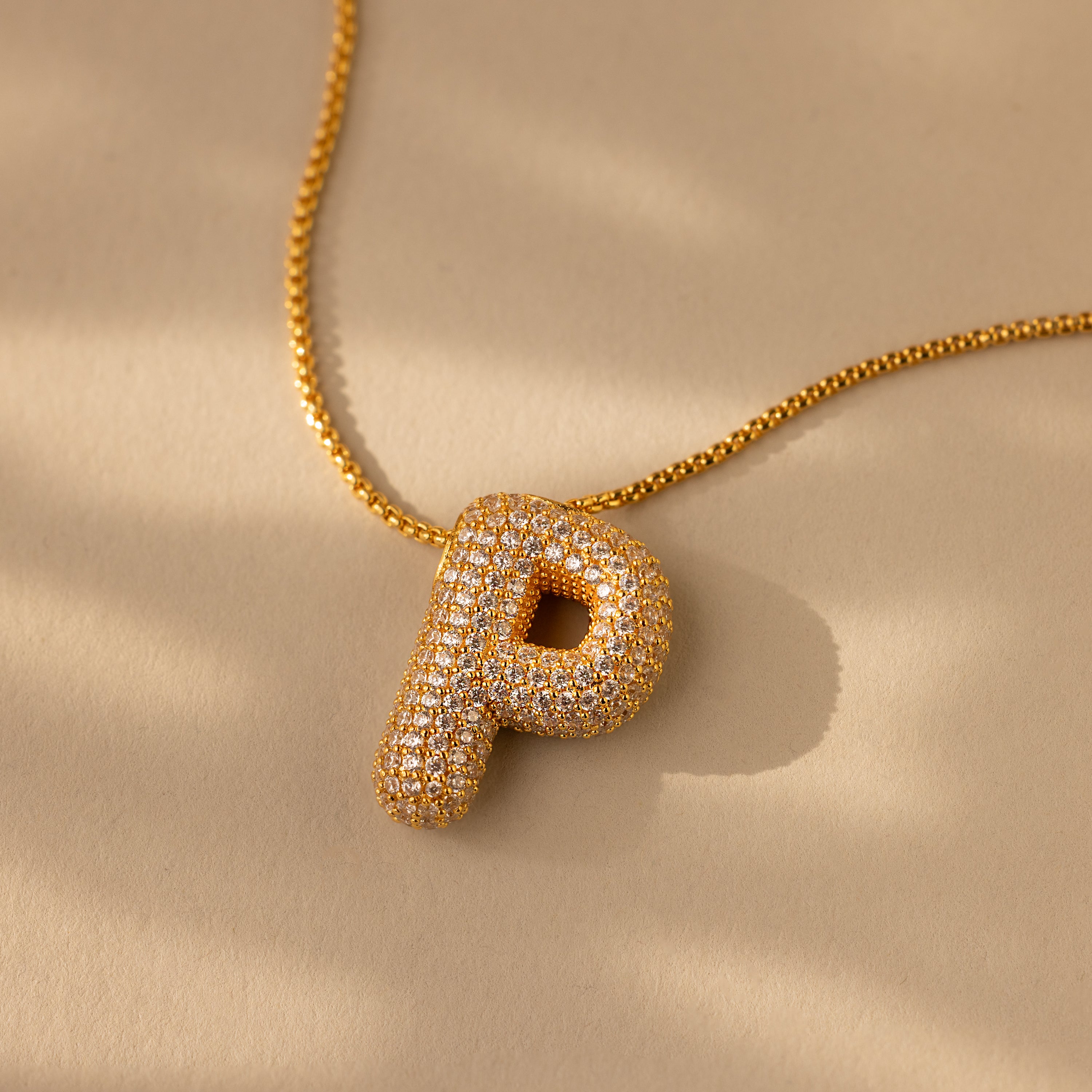 The Pave Puff Letter Necklace in 18K Gold features a gold "P" letter charm with sparkling pave diamonds, elegantly displayed on a beige background.