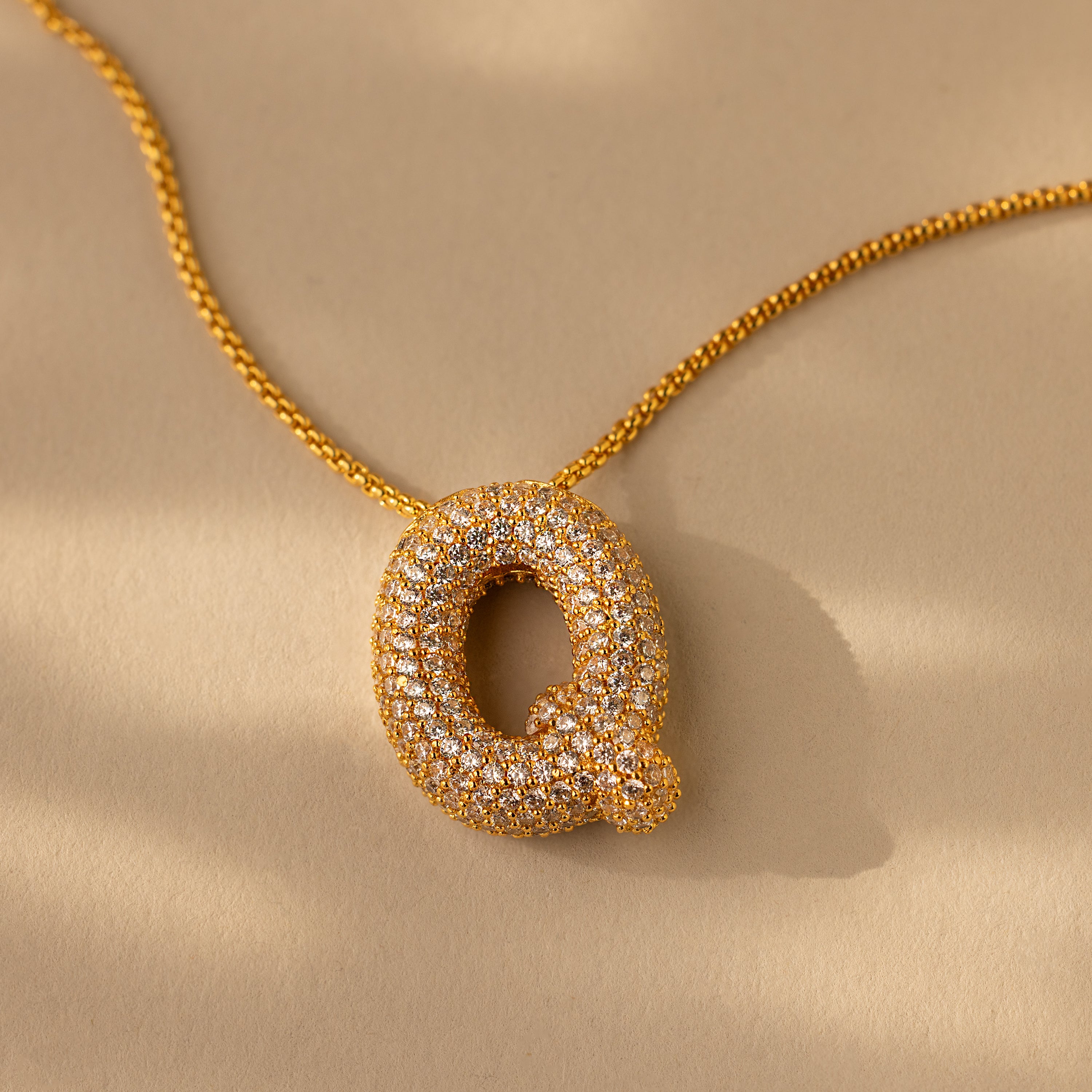 The Pave Puff Letter Necklace in 18K Gold features a diamond-encrusted Q pendant on a beige background, adding a timeless, personalized sparkle with its elegant gold letter charm.