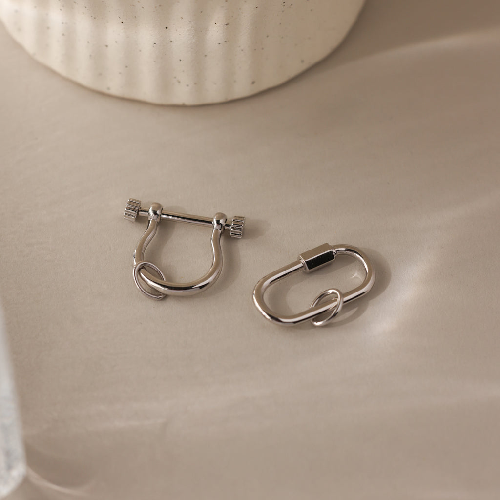 The Bar & Carabiner Charm Set features two silver ear cuffs with geometric designs on a beige surface—ideal as charm connectors or for pairing with favorite necklace accessories.