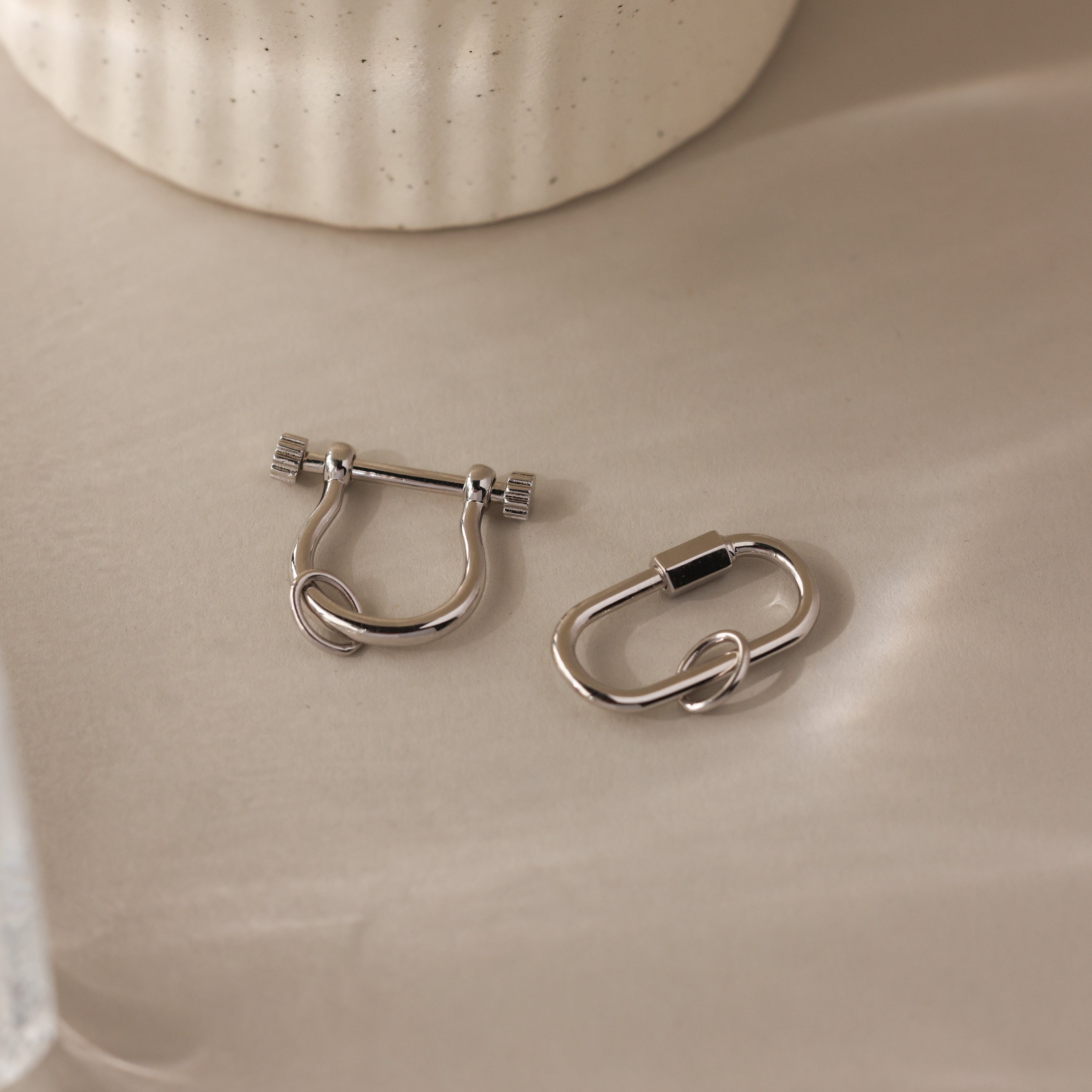 The Bar & Carabiner Charm Set features two silver ear cuffs with geometric designs on a beige surface—ideal as charm connectors or for pairing with favorite necklace accessories.