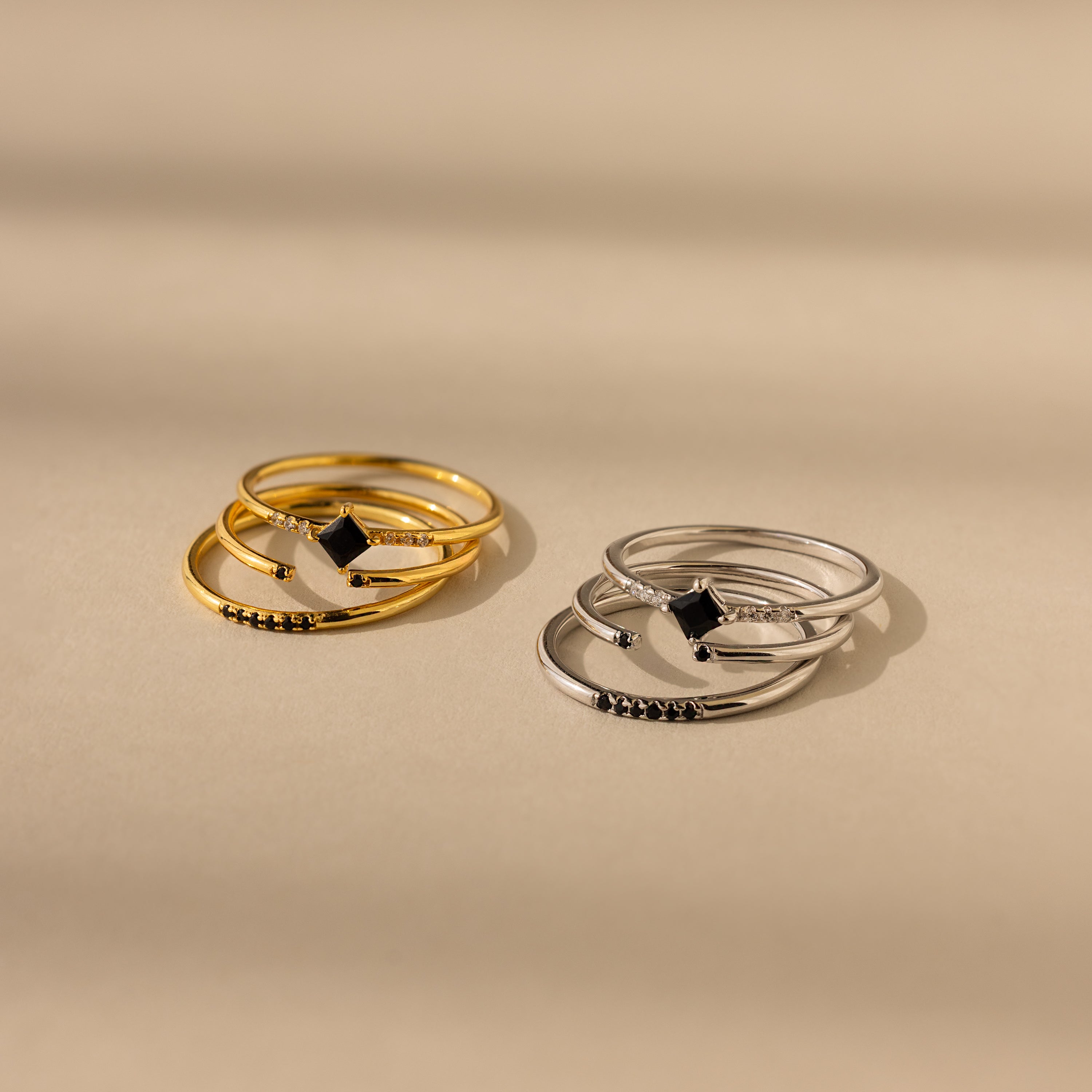 Two sets of stacked rings, one gold and the Black Abyss Ring Set in Silver, feature dainty bands with geometric and heart-shaped designs. The Black Abyss set showcases sleek black gemstones on a beige surface for a striking look.