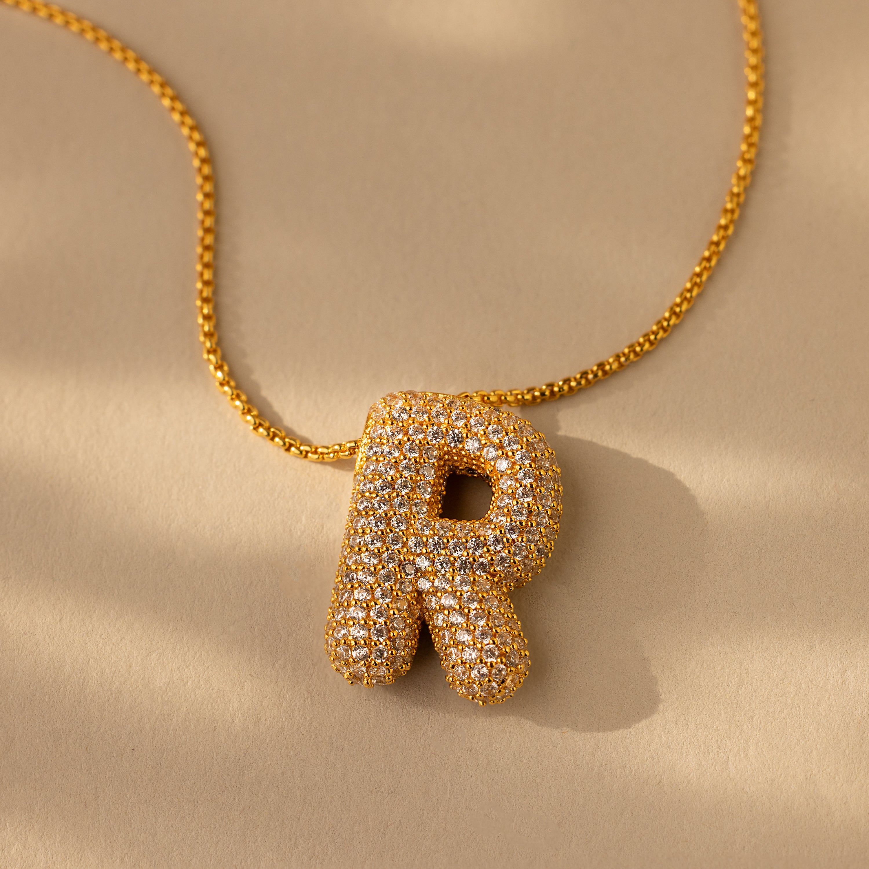 The Pave Puff Letter Necklace in 18K Gold features a large, sparkling pave puff letter R pendant encrusted with clear stones against a beige background.