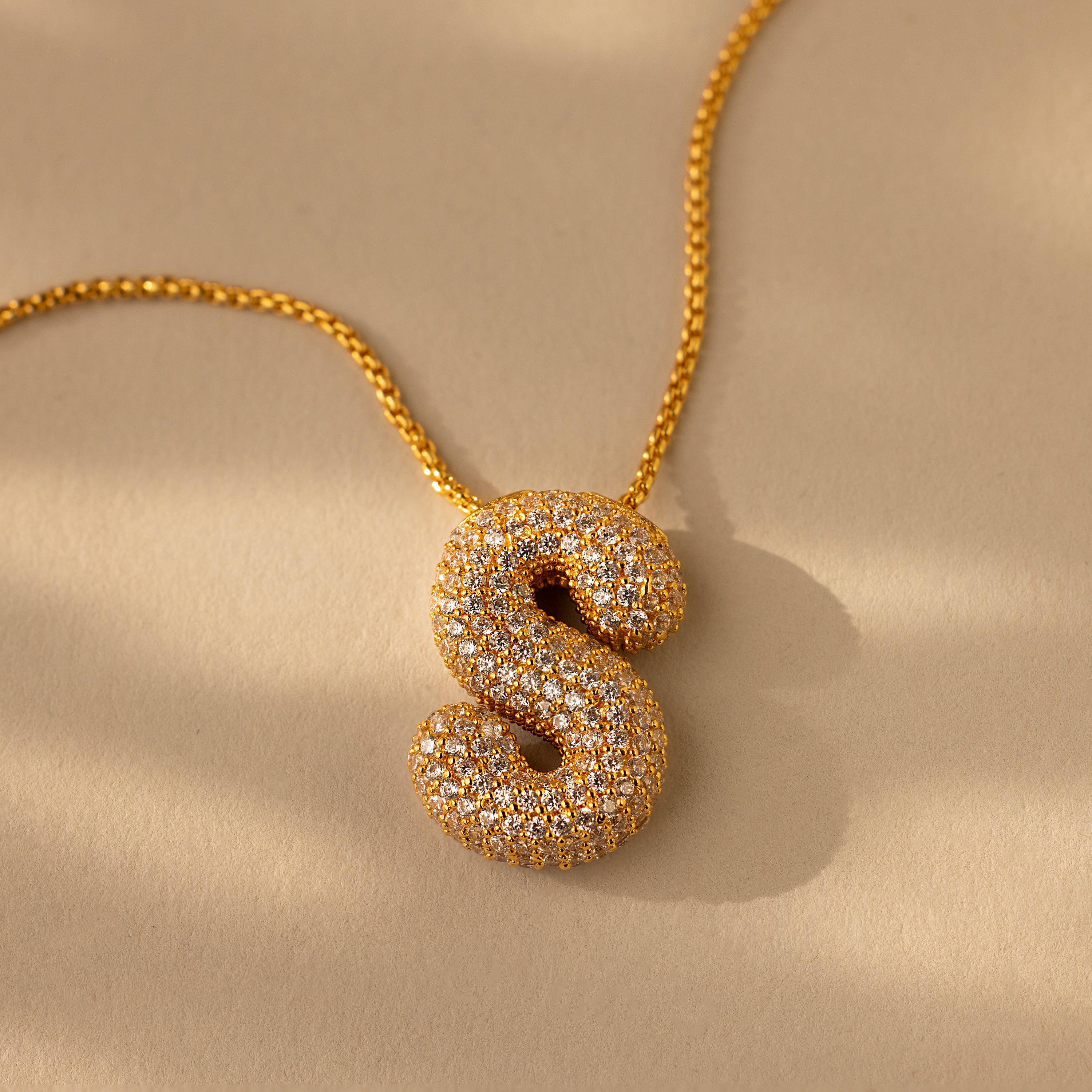 The Pave Puff Letter Necklace in 18K Gold features a diamond-studded S-shaped pendant, shown on a beige background.