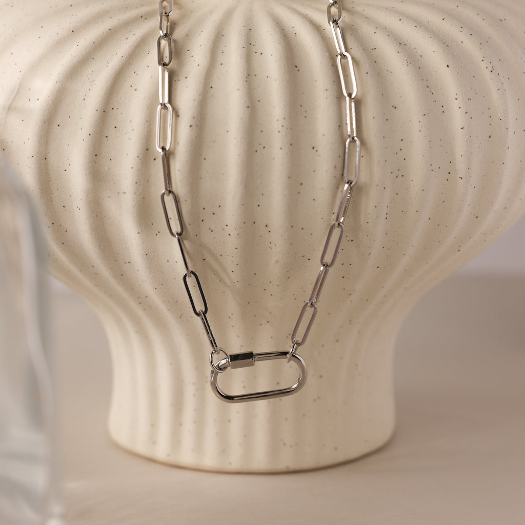 The Bar & Carabiner Charm Set in White Gold features a chunky chain necklace with a large carabiner clasp, displayed on a textured cream-colored vase.
