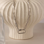 The Bar & Carabiner Charm Set in White Gold features a chunky chain necklace with a large carabiner clasp, displayed on a textured cream-colored vase.