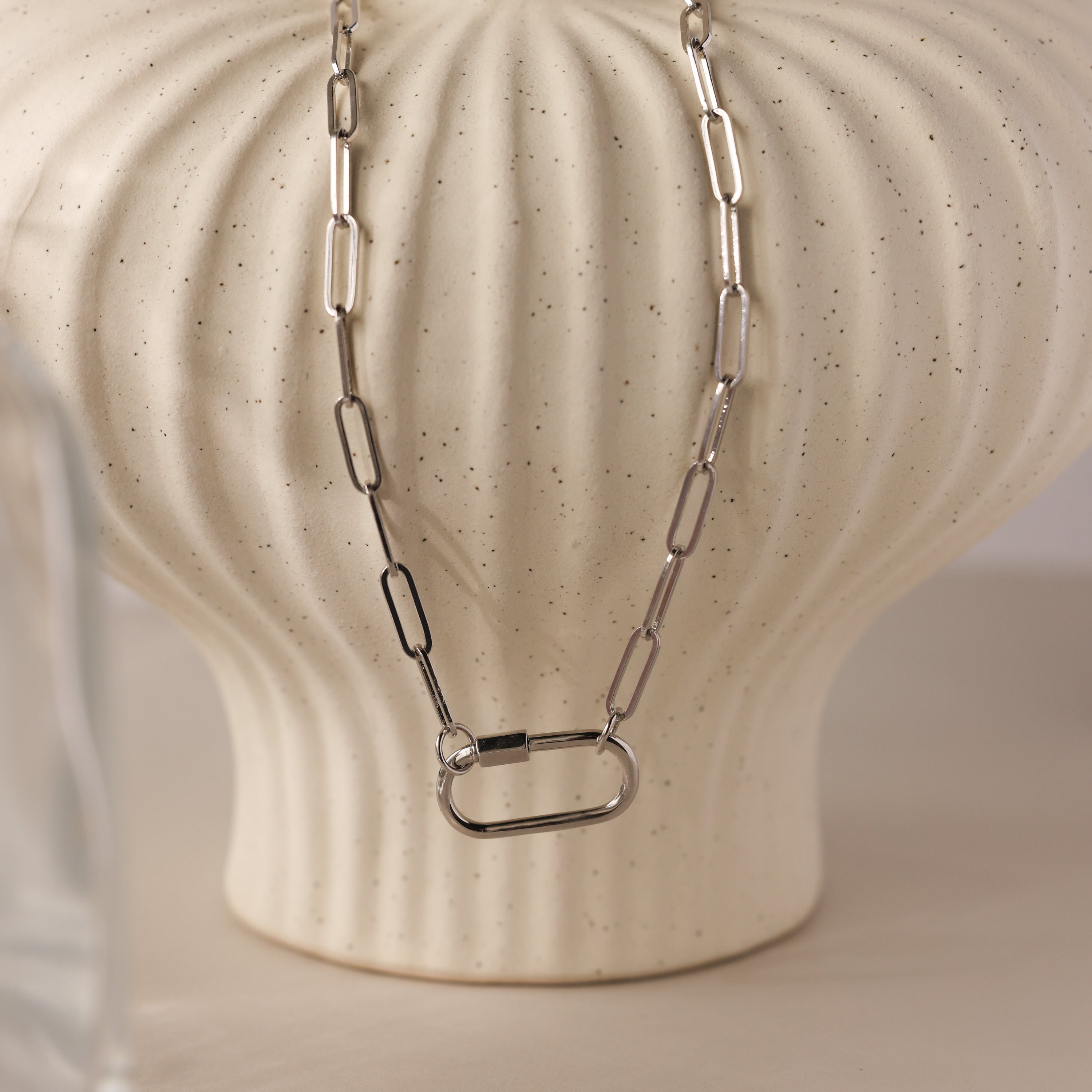 The Bar & Carabiner Charm Set in White Gold features a chunky chain necklace with a large carabiner clasp, displayed on a textured cream-colored vase.